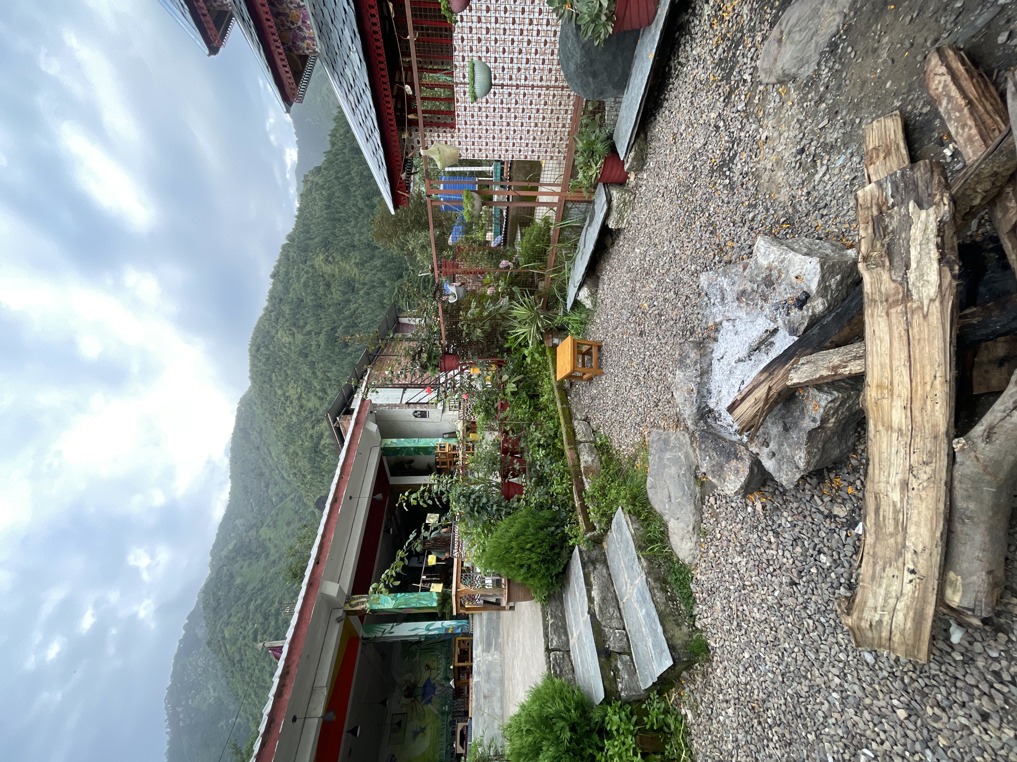 In Haini, India, this image captures an outdoor seating area and entrance to a building. In the foreground, logs of wood are stacked in front of a fire pit constructed with large stones. The ground is covered with small grey pebbles. To the left, a set of stone steps leads up to a building with an open-air entrance and red trim. The building's interior is visible, with tables and chairs set up, and a large mural on the back wall. A lush garden with various plants and potted flowers borders the entrance. To the right, a similar building with a red roof and decorative trim is visible, featuring a patterned tiled wall and hanging planters. The background is dominated by a steep, heavily forested mountain range under a cloudy sky.