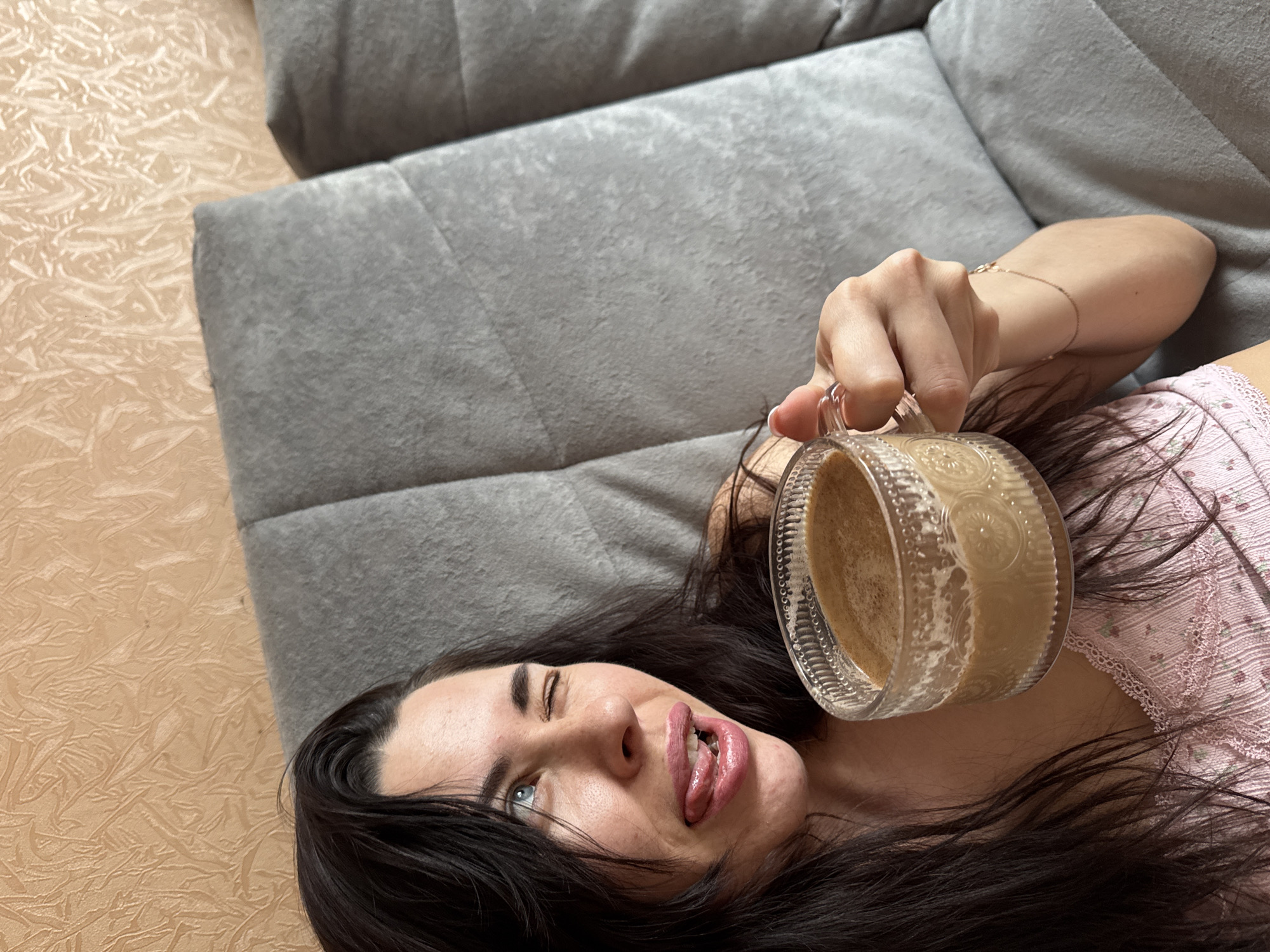 A woman with long, dark hair and blue eyes is lying on a gray couch. She is wearing a light pink top with floral patterns. Her right eye is closed in a wink, and her tongue is sticking out. She is holding a clear, decorative glass mug filled with a light-colored beverage. Her right arm is extended, holding the mug above her. A thin, gold bracelet is visible on her wrist. The location is the city of Chaikovsky, Russia.