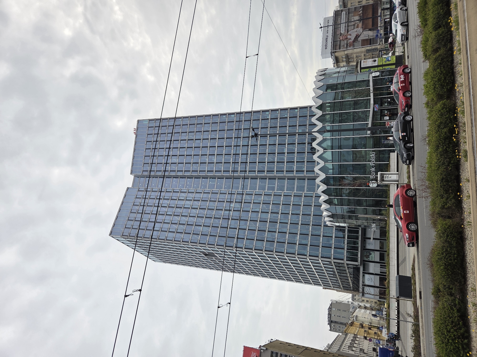Here is a description of the image:
The image captures a view of a modern glass-fronted building in Warsaw, Poland, identified by the "Bank Polski" signage on its facade. The building's design features a zigzag-patterned architectural element near the top of the lower section, creating a distinctive aesthetic. Adjacent to the building is a street with a line of cars parked along the curb.
Among the vehicles are two cars marked with "Bolt," suggesting they are part of a ride-sharing service. The street is bordered by a narrow strip of greenery, featuring shrubs and yellow flowers. Above the building, overhead tram lines extend across the frame, adding a layer of urban infrastructure. In the background, other buildings and billboard advertisements are visible. The sky is overcast, contributing to the soft lighting of the scene.