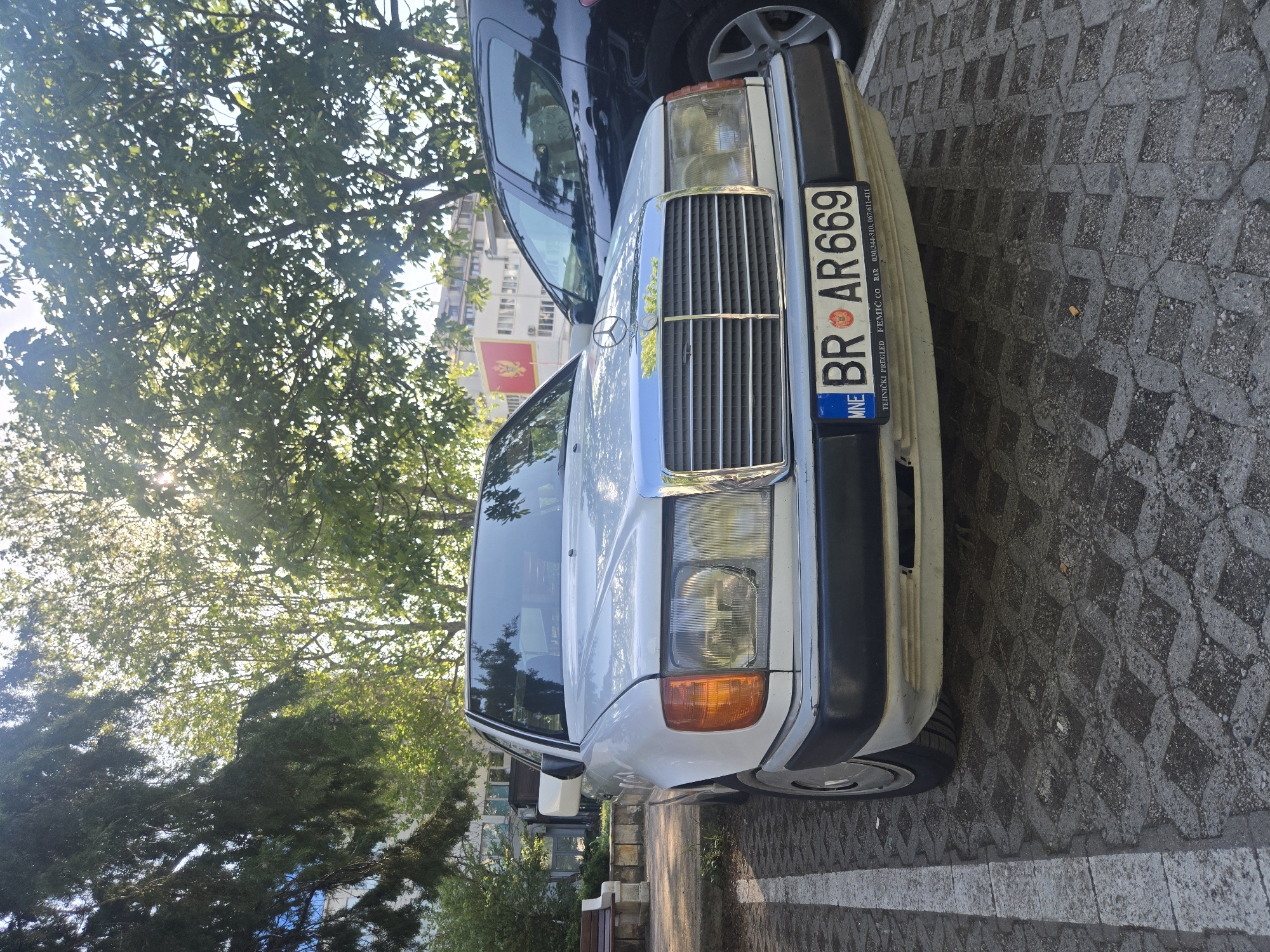 The image shows the front of a white Mercedes-Benz sedan parked on a paved surface. The car has a Montenegro license plate reading "BR AR669" along with the details "TEHNICKI PREGLED FEMIC CO BAR 030/344-310, 067/611-411". A black SUV is partially visible behind it. The background includes trees with sunlight filtering through, and a building with a Montenegrin flag on top can be seen in the distance. The location appears to be an urban street in Bar, Montenegro. A bench is visible on the side.