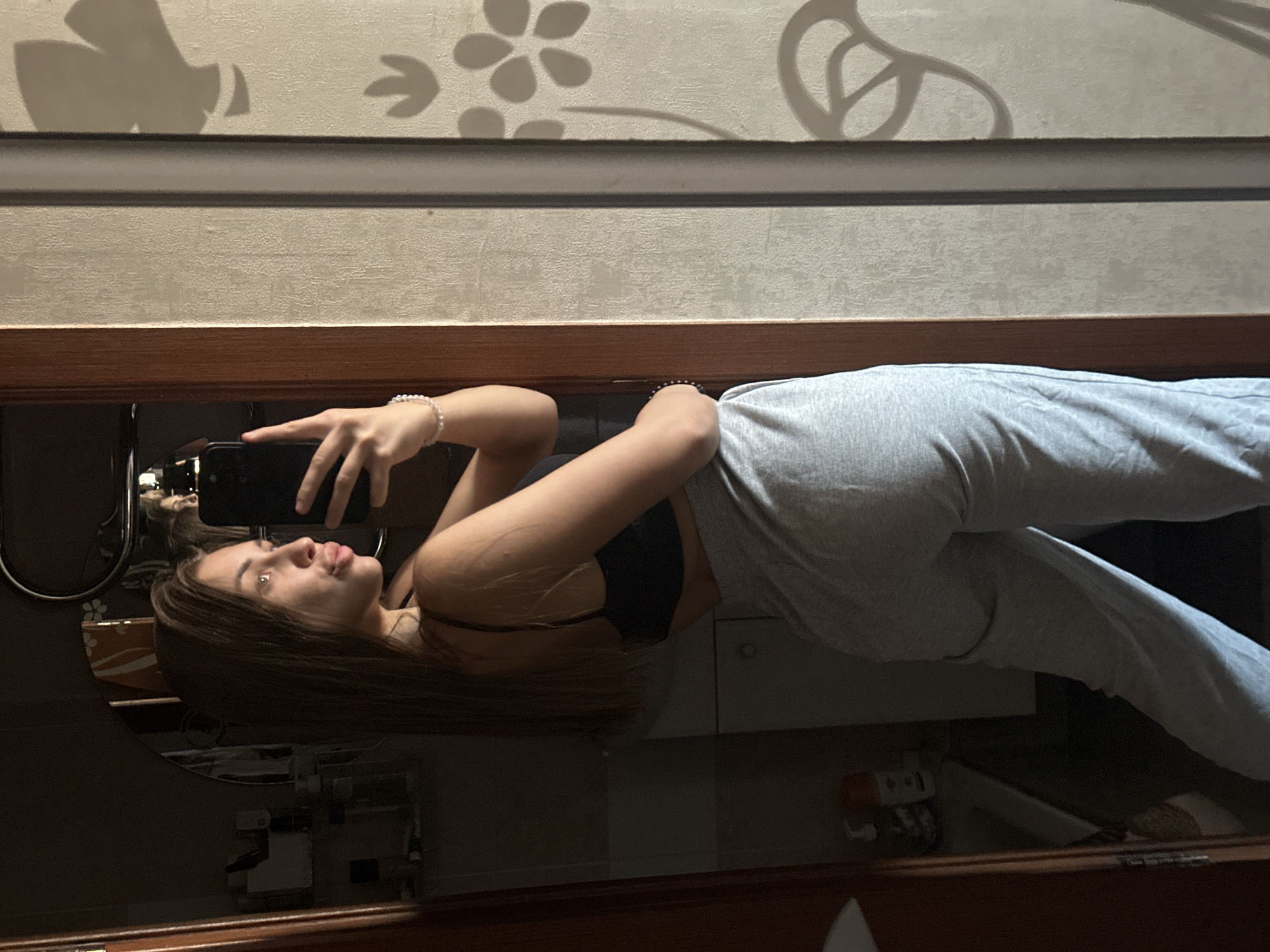 A young woman poses for a selfie in a bathroom mirror. She is wearing a black top and gray sweatpants. The background appears to be a bathroom interior with a vanity and patterned wallpaper. The location in the city of Irkutsk, Russia, is not confirmed by visual information.