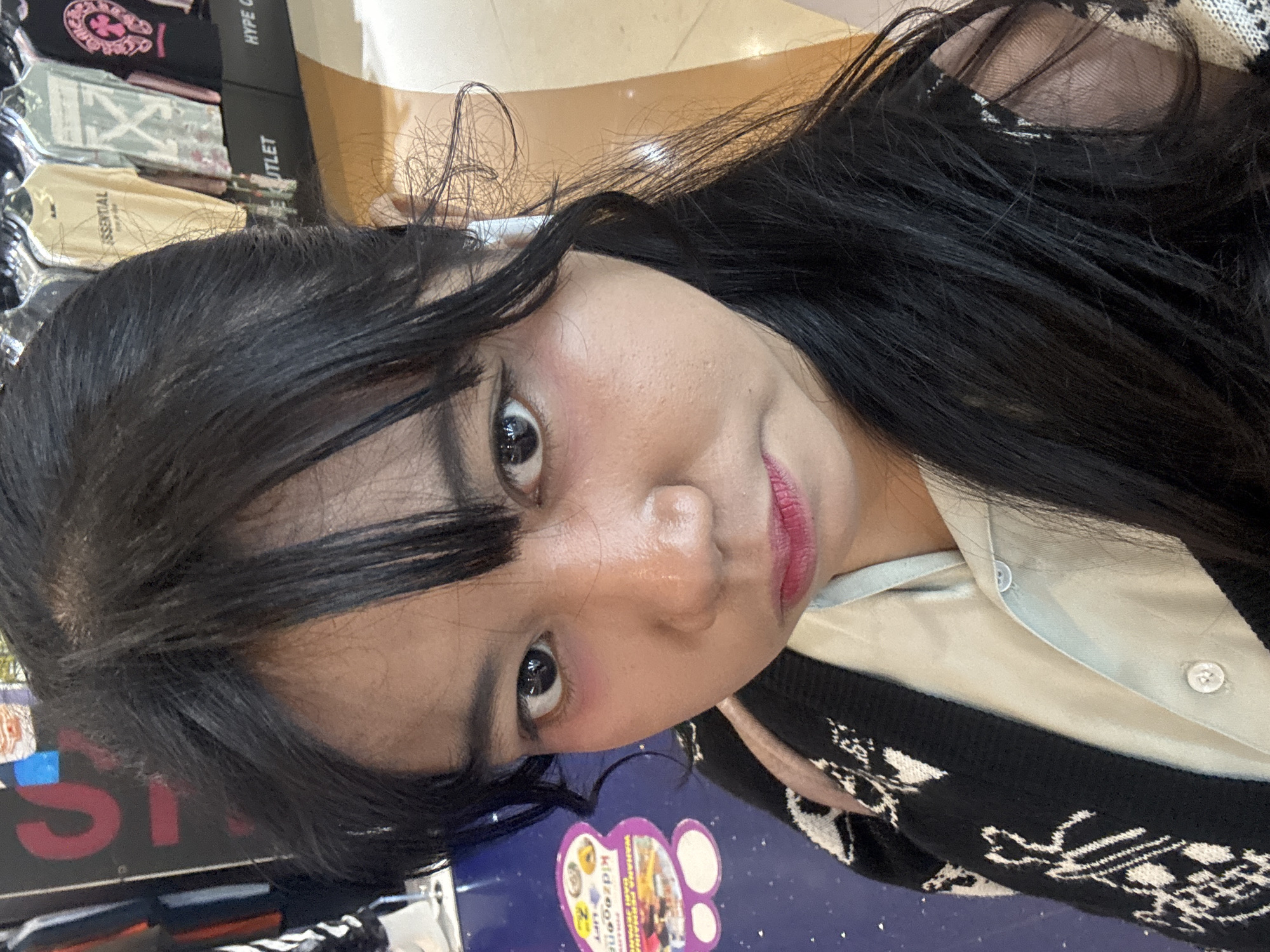 A young woman with dark hair and bangs is taking a selfie in what appears to be a shopping mall in North Jakarta, Indonesia. She is wearing a black cardigan with a white pattern over a light blue collared shirt. Her makeup includes pink blush and lipstick. In the background, there are racks of t-shirts for sale with brands like "ESSENTIAL" and "HYPE OUTLET" visible. A portion of a blue table with stickers is in the foreground to the left.