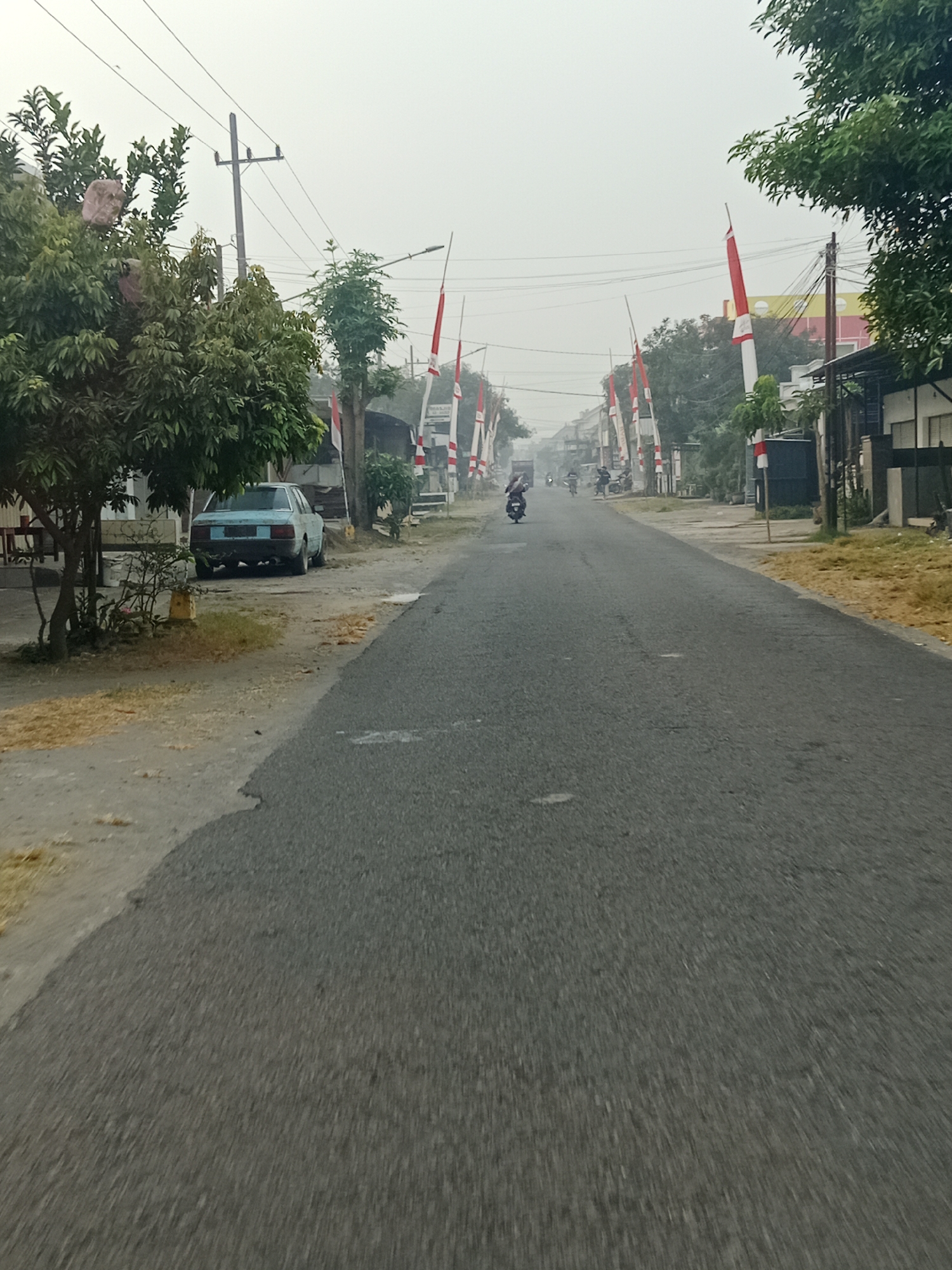 In the city of Srikaton, Indonesia, a road is depicted with a hazy sky. The road is asphalt and appears to be a two-lane street. On the left side of the road, a light blue car is parked on a dirt shoulder adjacent to a tree. Further down the road, several people are riding motorcycles. The sides of the road are lined with red and white flags, which are commonly seen during Indonesian Independence Day celebrations. Some buildings are visible on both sides of the street, and utility poles with wires are present. The overall scene suggests a residential or commercial area experiencing a slight atmospheric haze.