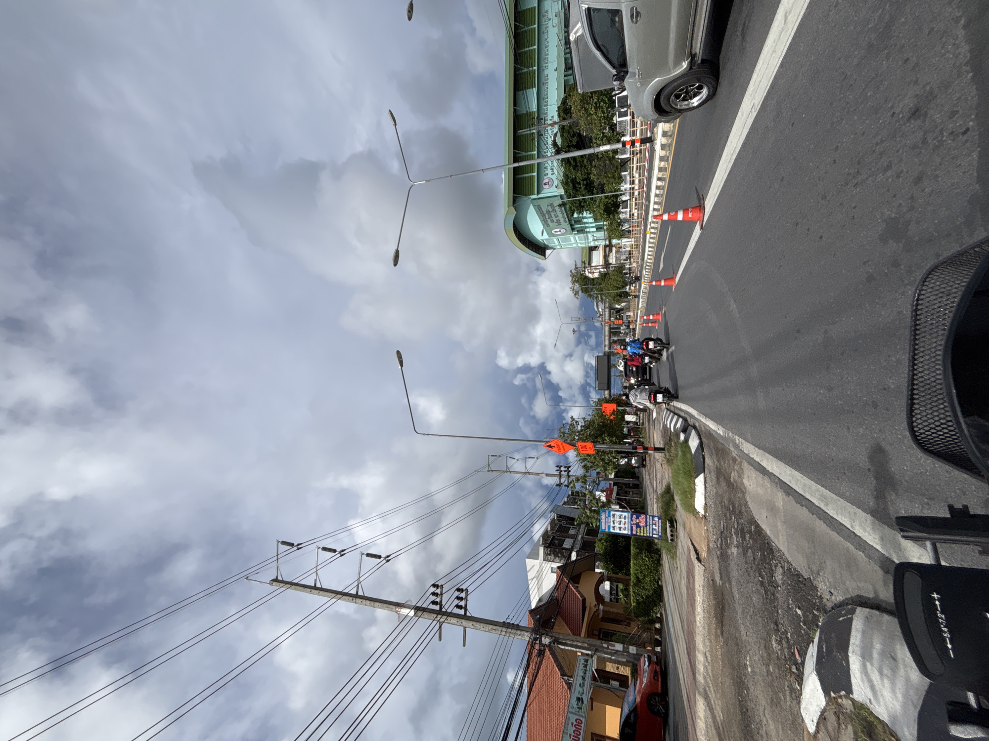The image depicts a street scene in Rawai, Thailand, under a cloudy sky. The perspective is from the viewpoint of someone riding a bicycle, as indicated by the handlebars and basket visible in the foreground. The road is paved and appears to be a multi-lane street with traffic.
On the left side of the road, there are buildings with orange-tiled roofs, some with signs indicating businesses. A red car is parked alongside the road. Utility poles with numerous electrical wires are a prominent feature along the left side of the street.
The road itself has traffic, including several motorcycles and a silver pickup truck. Orange traffic cones are placed along the right side of the road, suggesting some form of roadwork or traffic management. Tall streetlights are also visible, spaced along the road.
On the right side of the street, further down, is a large, teal-colored building with a curved roof, likely a public facility or school, with signage in Thai. Trees and greenery are present along the sidewalks and in front of the buildings. The overall environment appears to be a moderately busy urban or suburban area.