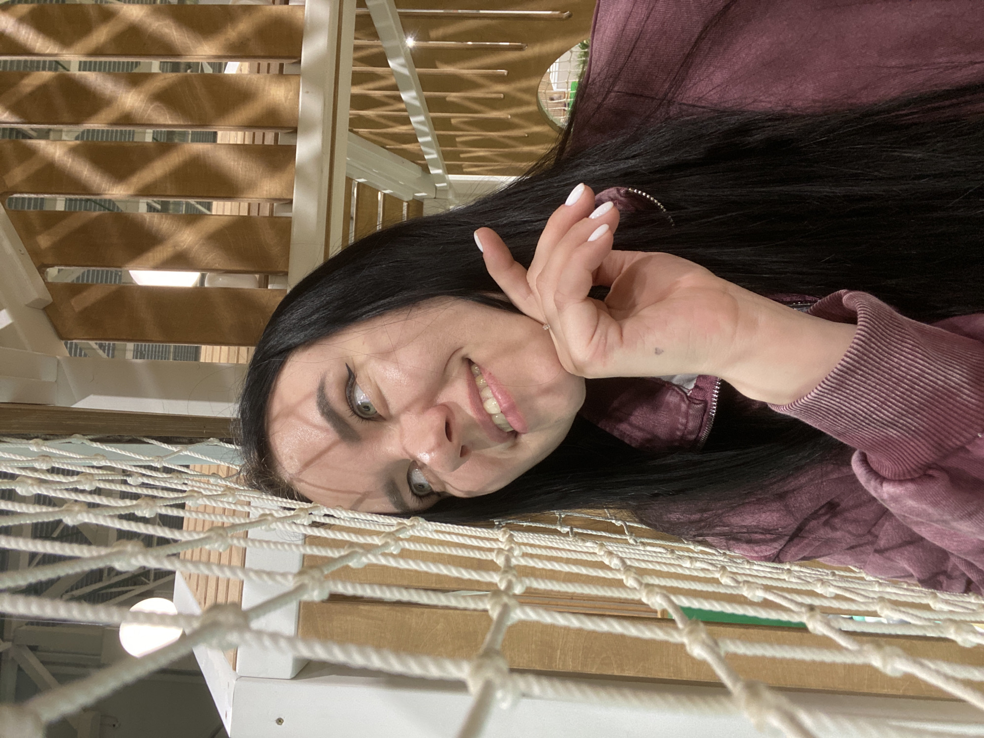 A person is lying on a net indoors, possibly in a recreational area. The person has dark hair, fair skin, and is wearing a pink or maroon zippered jacket. The person is smiling and making a hand gesture. The background includes wooden structures and a light fixture. There is no verifiable evidence to confirm the location in the city of Perm, Russia.
