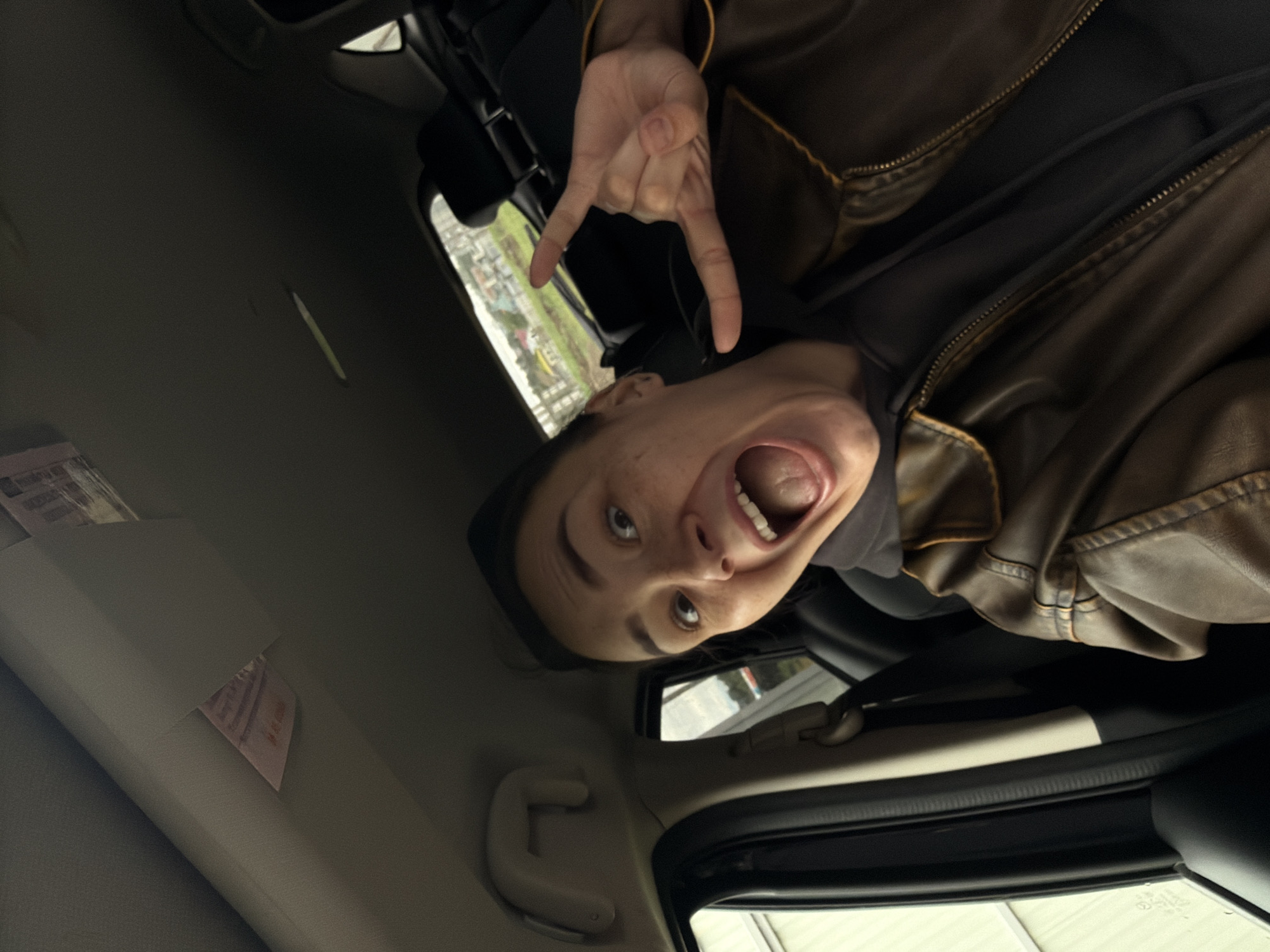 A person with a leather jacket is in a car, making a "rock on" hand gesture. The car interior is visible, including the dashboard and interior ceiling. Outside the window, buildings suggest a residential area. The exact location in Chaikovsky, Russia, cannot be determined from the image.