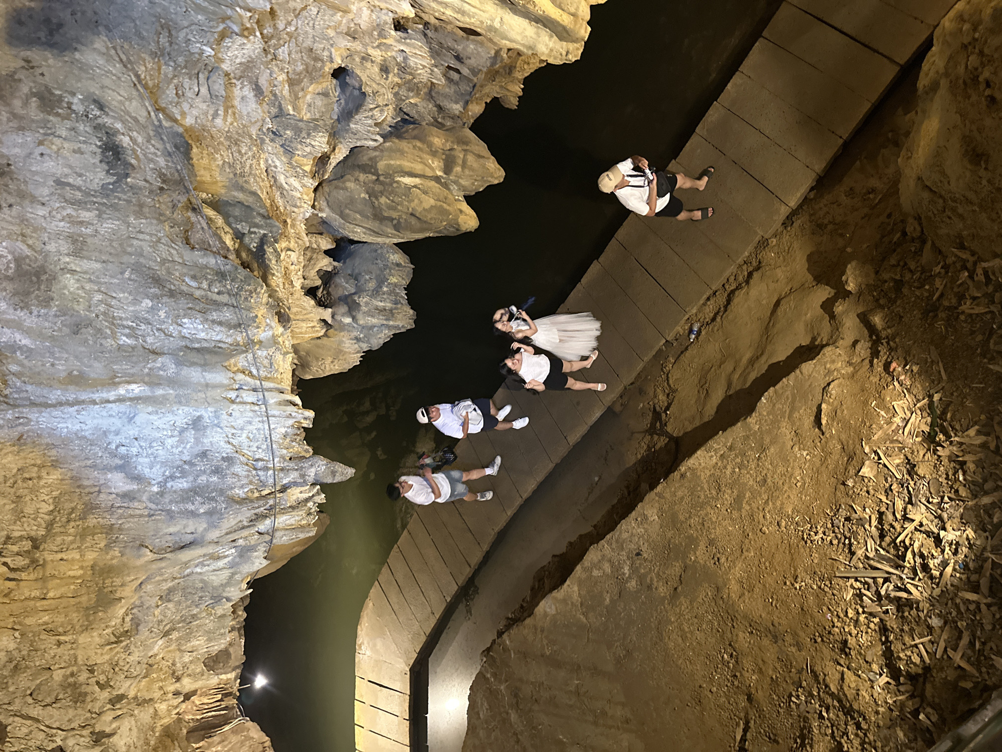 The image depicts a group of tourists walking on a wooden pathway inside a cave, illuminated by artificial lights. The cave features impressive stalactites and stalagmites, with dark water flowing alongside the path. The pathway is constructed from wooden planks and is positioned above the water. Several individuals are visible, some walking and others posing for photos. One person, a woman in a white dress, appears to be posing with two other women. Another person, a man wearing a hat, is holding a camera and looking at it. The environment is dimly lit, with the cave walls casting shadows. The overall scene suggests a tourist attraction within a natural cave setting. The location is identified as Yên Sơn Ward, Vietnam.