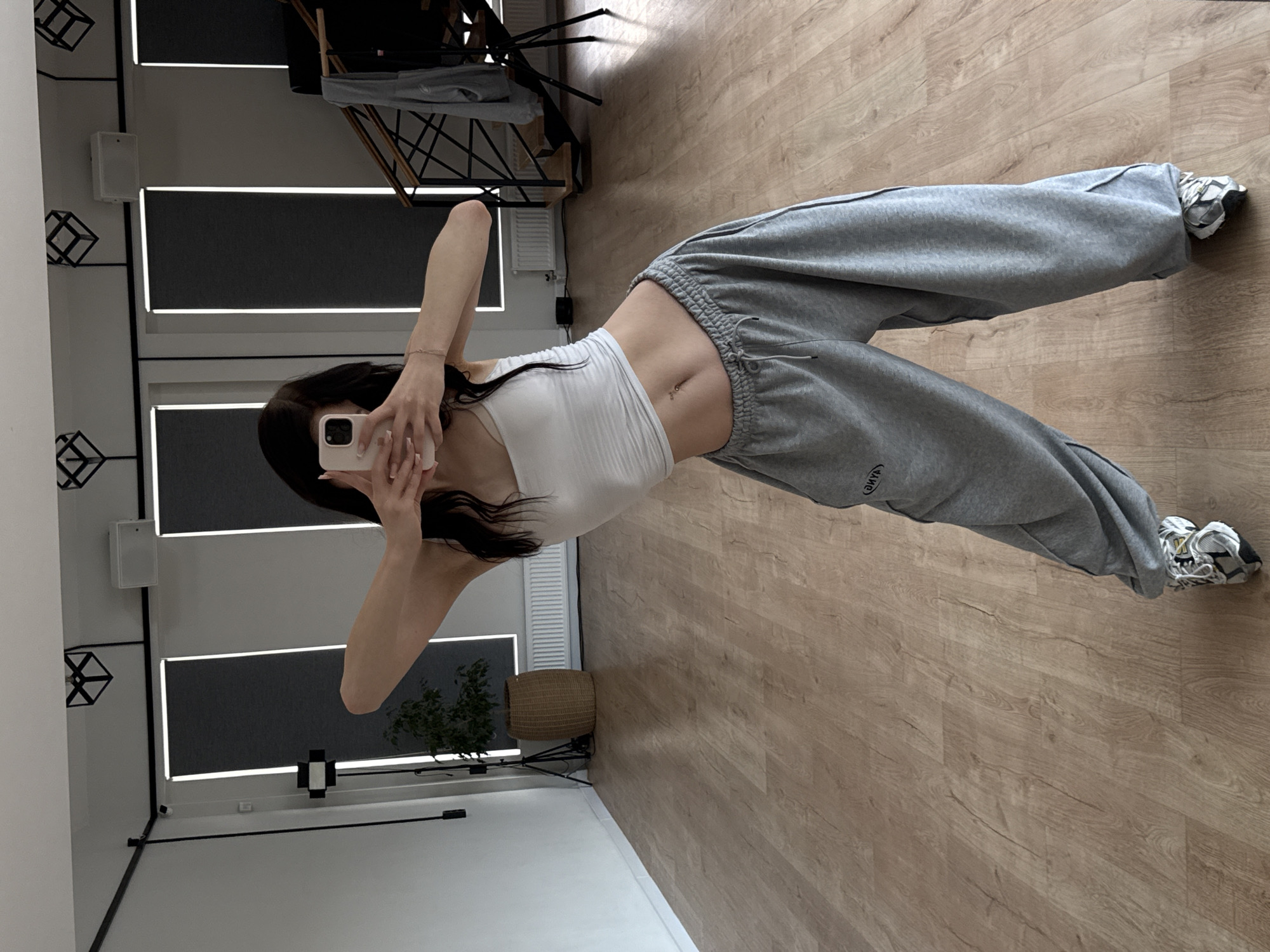A full-body shot captures a young woman taking a mirror selfie in what appears to be a home gym or studio. She is standing, with her body angled slightly away from the camera, and holding her phone up to take the picture. She is wearing a white crop top that exposes her midriff and grey sweatpants. The sweatpants are loose-fitting and appear to have some text or a logo on the left leg, though it is partially obscured and reversed due to the mirror. She is also wearing athletic shoes.
The setting is a room with light brown wood flooring and white walls. In the background, visible in the mirror's reflection, are several features: a tall, dark metal rack with clothing hanging on it, several rectangular decorative elements on the wall, and blinds or shutters on a window. There is also a small potted plant and a woven basket. The lighting suggests it is daytime, with natural light likely coming from the window. The overall mood is casual and relaxed, typical of a personal photo session. The location is identified as Zakharovo, Russia.