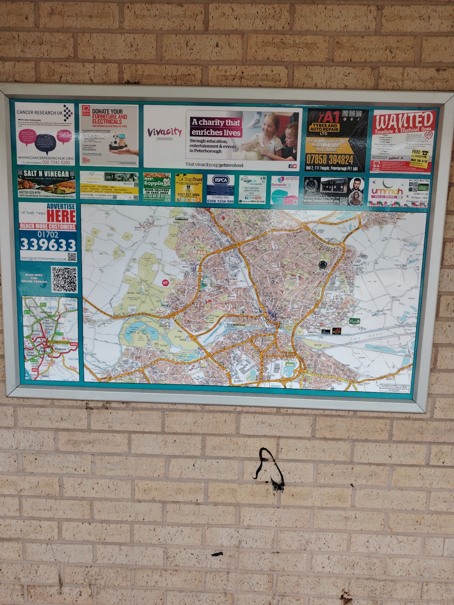 The image displays a large, rectangular public information board mounted on a light brown brick wall. The board features a framed map of Peterborough, United Kingdom, surrounded by multiple advertisements.
The main subject of the board is a detailed street map of Peterborough and its surrounding areas, including towns and villages like Werrington, Orton Wistow, Stanground, Bretton, and Gunthorpe. Major roads such as the A1, A15, and A605 are clearly marked. A prominent black circular marker is affixed to the map near the top center, pinpointing a specific location within Peterborough. The map also shows geographical features like Milton Park and various bodies of water. The map's bottom left corner includes a wider area context showing Peterborough relative to Bedford and Level.
Above and to the left of the map, there are several square and rectangular advertisements. From left to right in the top row, these include ads for "Cancer Research UK," a "Heart Foundation" appeal to "Donate Your Furniture And Electricals," "Vivacity presents A charity that enriches lives" (referencing educational and entertainment events in Peterborough), "A1 Tyres And Auto Repair Ltd" located at 111 Fengate, Peterborough PE1 5GA, and an ad for "WANTED Furniture & Electrical items."
Below the top row of ads and directly above the map, another row of smaller advertisements is visible. From left to right, these include "Salt N Vinegar" fish and chips (located at 12B3 Lincoln Road, Werrington, Peterborough PE4 6LJ), "Glisten" beauty treatments, "Mr. Poppings" home delivery, "Dogs Trust," and "RSPCA."
To the left of the map, a large blue box advertises "ADVERTISE HERE" with a phone number (01702 339633) and email address (info@uktownmaps.co.uk). Below this, a section titled "SCAN HERE FOR ONLINE VERSION" includes a QR code.
The entire display is enclosed in a metallic-looking frame with a turquoise inner border. The brick wall on which the board is mounted is composed of light tan or yellowish bricks with gray mortar lines. Below the board, towards the bottom left of the frame, a distinct black graffiti mark, abstract in form, is visible on the brickwork. The wall also exhibits some minor stains and discoloration. No people are present in the scene.