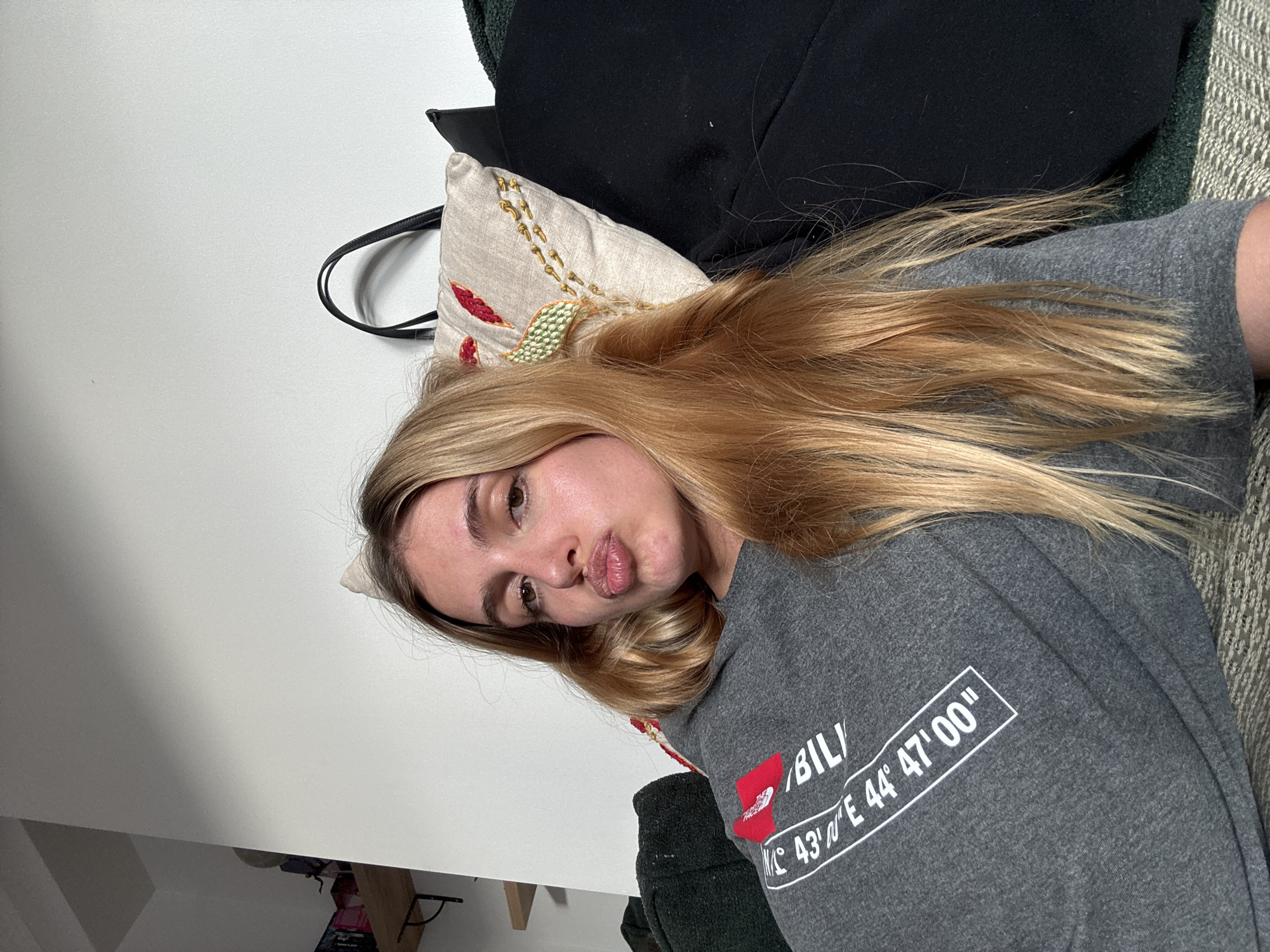 A woman with blonde hair is lying on a patterned gray couch, wearing a gray t-shirt with a red logo and the text "IBIL" followed by "N 43° E 44° 47' 00"". She is pouting her lips toward the camera. Her head rests on a small decorative pillow with red, green, and gold embroidery. Behind her are a black jacket and a green fleece blanket. The background shows a white wall on the left, and a shelf with items on the bottom left corner. The location in the city is Unknown, Unknown.