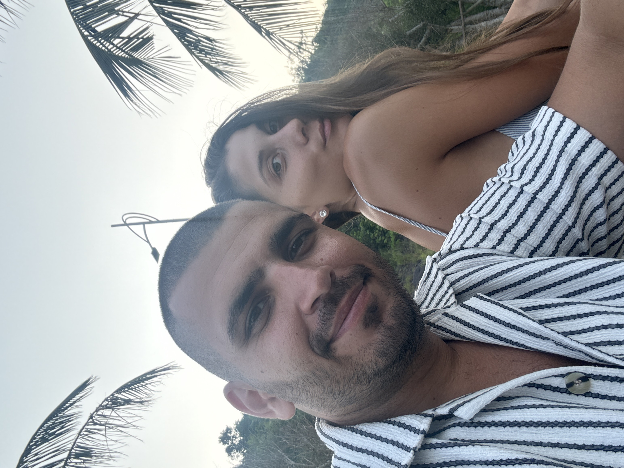 A couple poses for a selfie against a backdrop of palm trees and a pale sky, likely at dusk. The man, in the foreground, has short dark hair, stubble, and wears a striped black and white shirt. Behind him and slightly above, a woman with long brown hair looks at the camera. She is wearing a striped outfit with thin straps. Palm fronds are visible in the upper left and lower left corners of the image. A pole can be seen in the background. The exact location within Đà Nẵng, Vietnam is not discernible from the image.