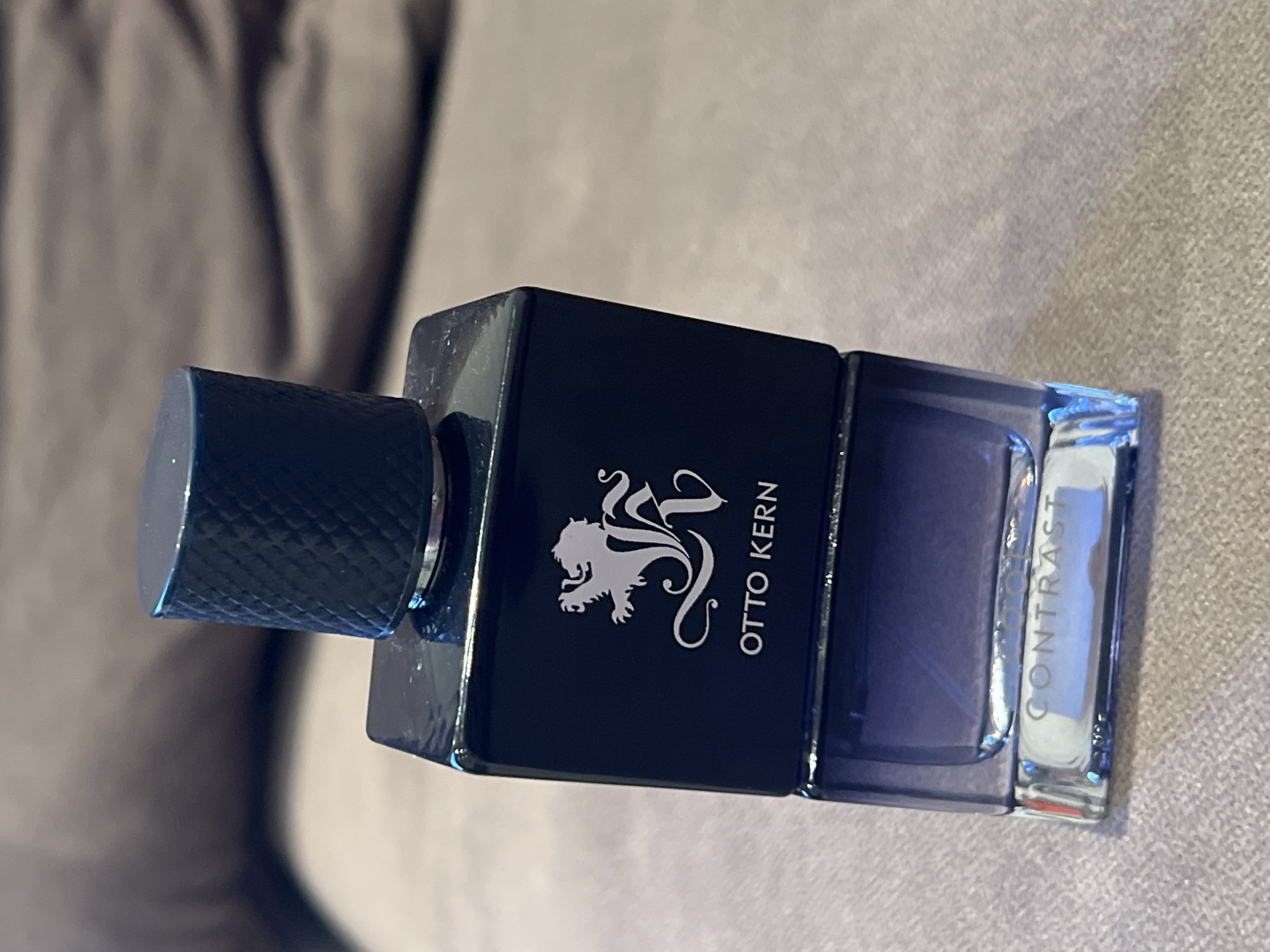 The image shows a close-up, eye-level shot of an "Otto Kern Contrast" perfume bottle. The bottle is dark blue and has a silver cap with a textured pattern. The brand name "OTTO KERN" and a stylized lion logo are visible on the bottle. The bottle appears to be sitting on a textured gray surface, possibly a sofa or upholstery, with a blurred background of a darker, possibly brown, fabric. There is no indication of the city of Montfermeil, France, in the image or its context.