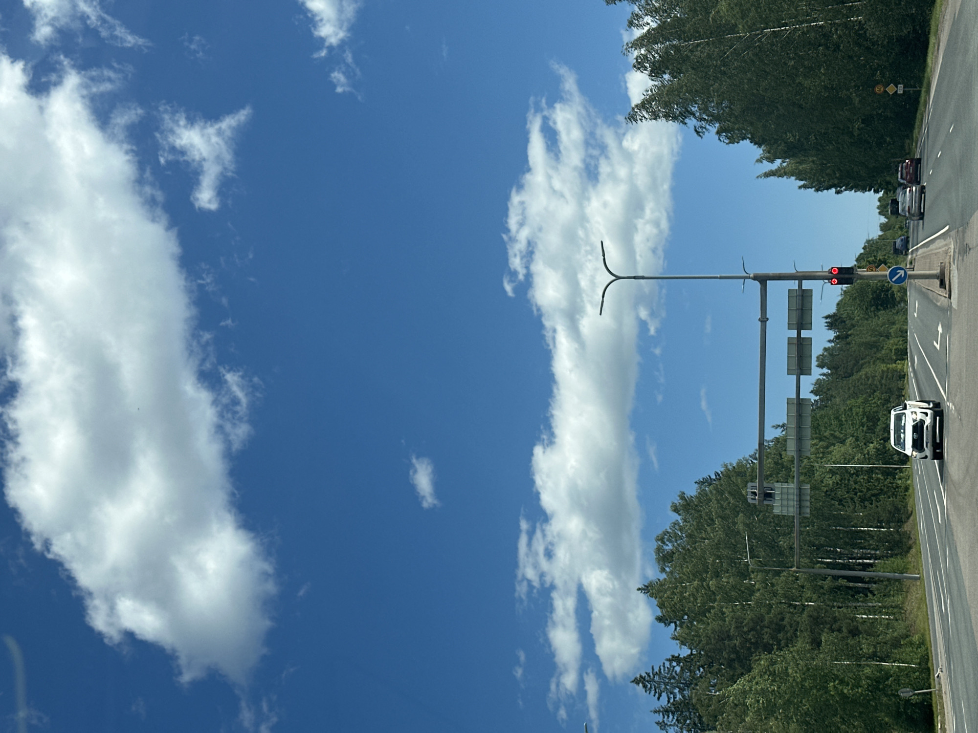 An outdoor, eye-level, medium shot captures a road scene on a sunny day. The sky is a vibrant blue with fluffy white clouds scattered across it. The road, paved with asphalt, stretches forward, bordered by lush green trees and vegetation on either side. A white SUV is driving on the road, and a traffic light displays a red signal. There are streetlights and road signs present. The scene appears to be in a suburban or rural area, likely outside of a city, given the abundance of trees and lack of prominent urban structures.
