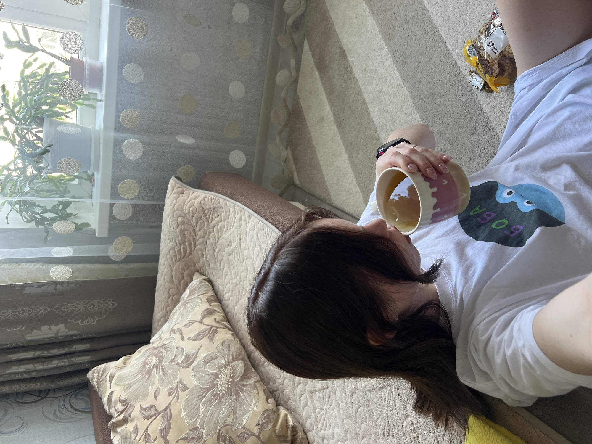 The image captures a high-angle view of a person lying on their left side on a light-colored couch or bed in an indoor setting.
**Subjects:**
The main subject is a young person with dark brown hair, seen from the back of their head and upper torso. They are wearing a white t-shirt. Their right arm is holding a mug, and a black wristwatch is visible on their left wrist. Their fingernails are painted light pink with small heart designs.
**Objects:**
The person holds a white ceramic mug with pink and purple wavy designs, filled with a light-colored liquid. The white t-shirt features a blue and black graphic design with the word "БОБА" (BOBA) written in green and pink letters. A yellow bag of snacks, identified as "ХРУМ - ХРУМ РИСОВЫЕ КРУПЫШКИ СО ВКУСОМ СЫРА" (Crunch-Crunch Rice Crisps With Cheese Flavor) and bearing the date "15.02.2023," is visible near their shoulder. The person is resting on a light beige couch or bed, which has patterned upholstery and a decorative cushion with a large floral design. In the background, a window is covered by a sheer white curtain adorned with textured circular patterns. Several potted plants, resembling a Schlumbergera (Christmas cactus), are placed on the windowsill. The floor or a large rug has a beige color with distinctive diagonal stripes.
**Activity:**
The person is in a relaxed, reclined position, holding a mug and appearing to be enjoying a beverage, suggesting a moment of leisure.
**Notable details:**
The scene is illuminated by bright natural light streaming through the window, indicating it is daytime. The overall atmosphere appears calm and comfortable. The Cyrillic text on the t-shirt and the snack packaging serves as a strong location cue, suggesting a Russian-speaking environment.
**Visible text:**
"БОБА" (BOBA) on the t-shirt. On the snack packaging: "ХРУМ - ХРУМ," "РИСОВЫЕ КРУПЫШКИ," "СО ВКУСОМ СЫРА," and "15.02.2023."