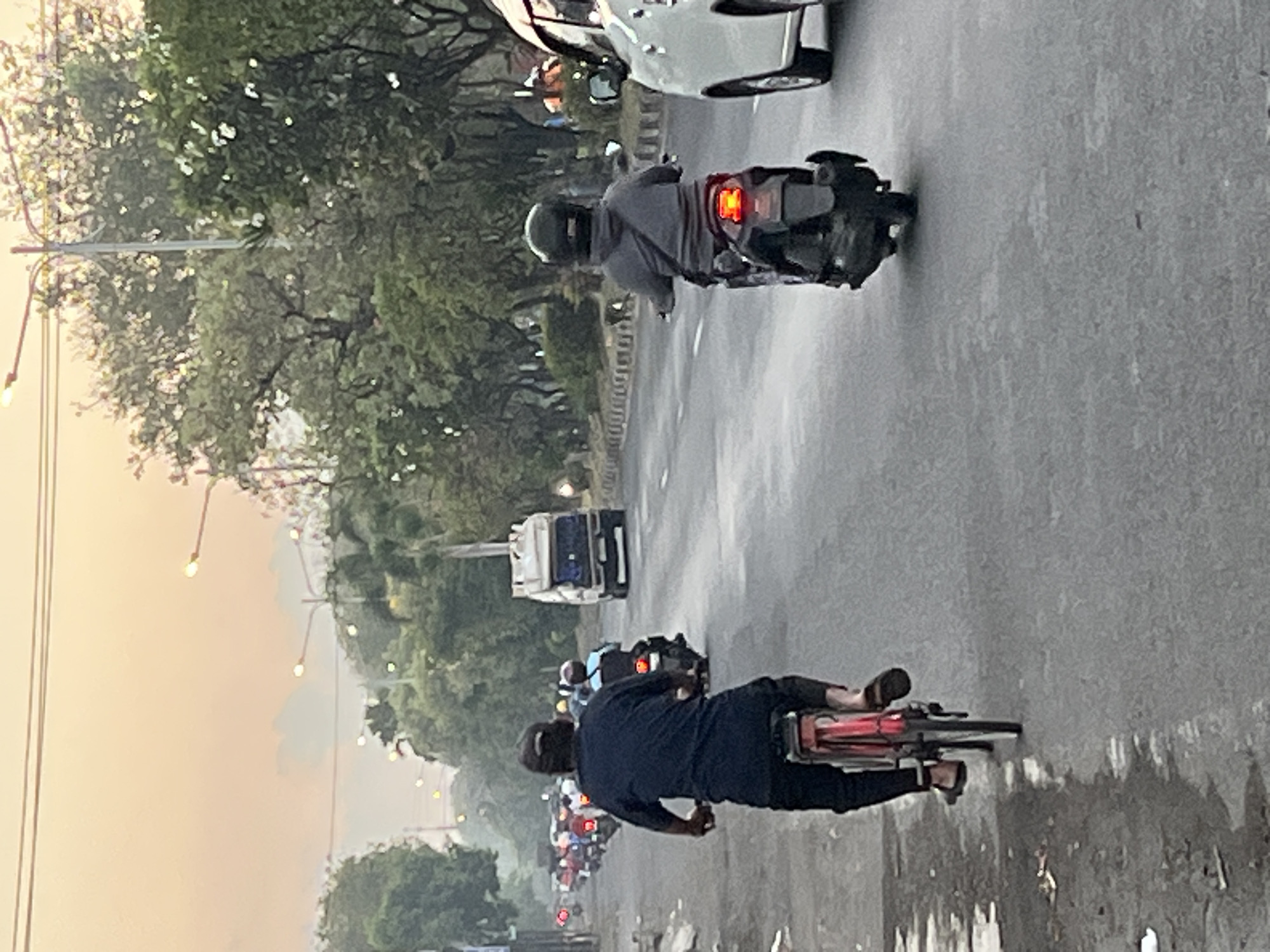 The image depicts a street scene in Ngaban, Indonesia, likely during dusk or dawn due to the soft, warm lighting and overcast sky. The foreground is dominated by a man on a bicycle, seen from behind. He is wearing a dark blue shirt, dark pants, a cap, and sandals. The bicycle is red. He is riding on the left side of the road.
Further down the road, several motorcycles are visible, also moving in the same direction. One motorcycle in the middle ground has a rider wearing a grey hoodie and a black helmet. To the right, a white car is passing by. In the distance, a white truck with a boxy cargo area is on the road.
The environment includes a paved road with markings, a sidewalk on the left with some debris, and a line of trees along the right side of the road. Streetlights are present, some illuminated, casting a gentle glow. The overall impression is one of moderate traffic on a city street.