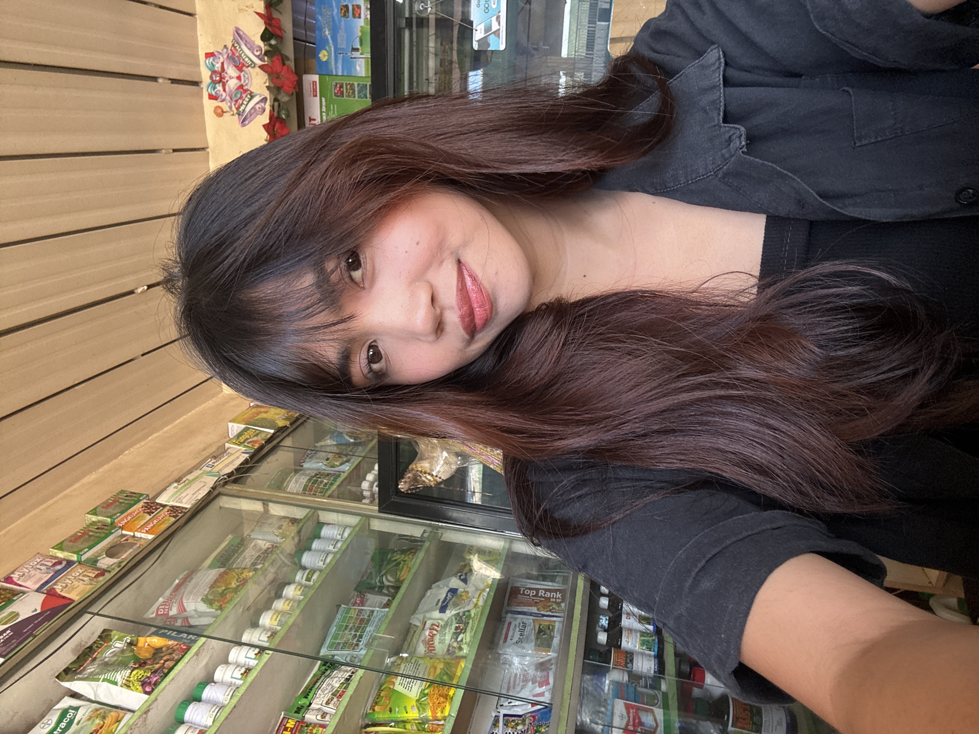 A young woman with long, dark brown hair and bangs smiles at the camera, her head tilted slightly to the left. She is wearing a black collared shirt over a black tank top. Behind her, shelves stocked with various products, likely agricultural supplies, are visible through a glass display. The shelves are filled with bags, bottles, and boxes, many of which have colorful labels. The lighting suggests an indoor environment, possibly a store. A festive Christmas decoration featuring bells and poinsettias is visible on the wall above the shelves. The location is identified as a store in Tagum City, Philippines.