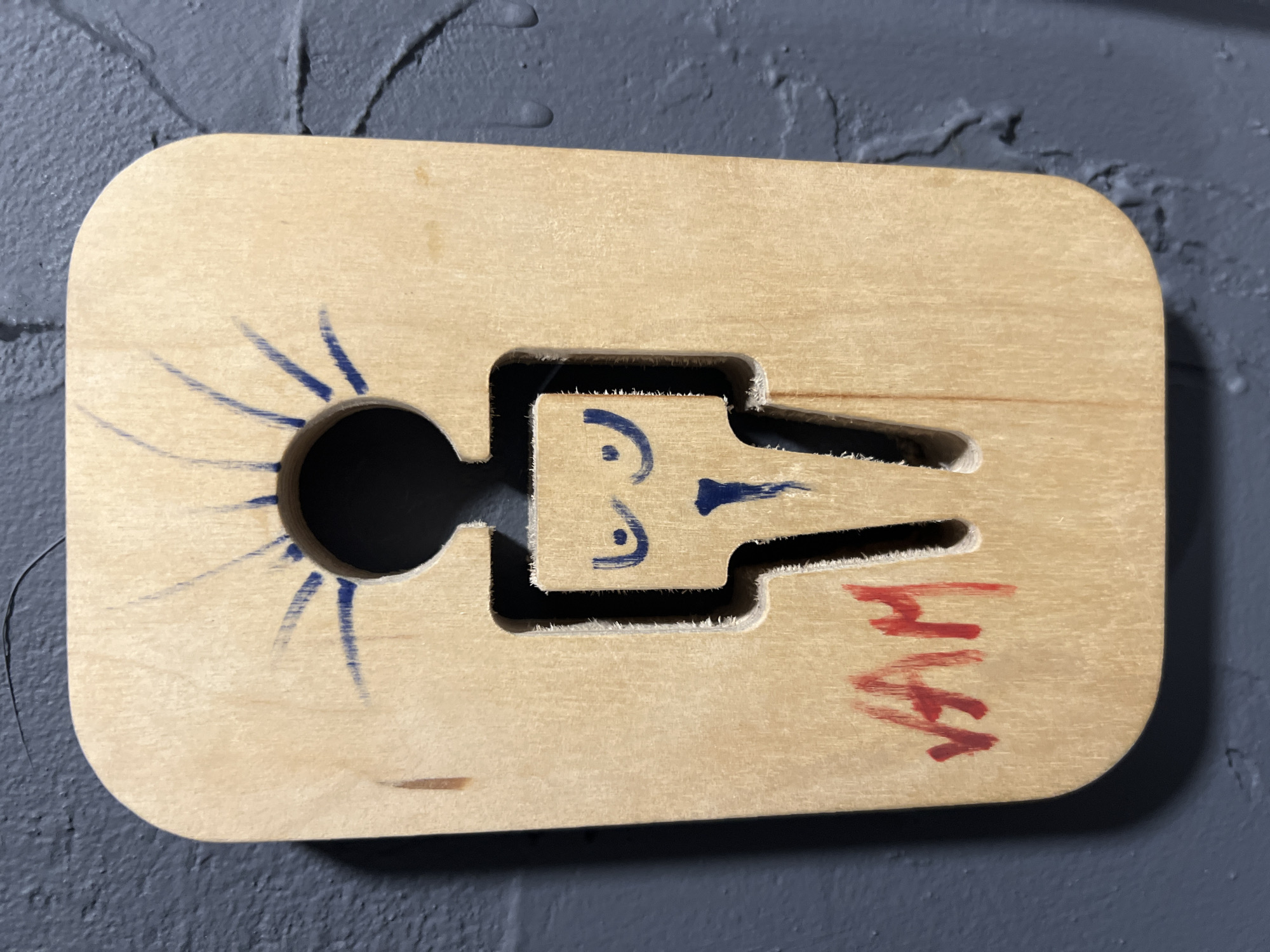 An image shows a wooden cutout of a female figure mounted on a dark grey textured wall. The cutout has a circular hole for the head, and the body is a stylized representation of a woman. Blue marker lines emanate from the headhole, resembling rays of light or a crown. Within the body cutout, blue marker lines depict breasts and a mouth-like shape. Red marker scribbles, possibly a signature or tag, are visible on the lower left of the wooden cutout, reading "VAM". The overall appearance suggests a handmade or artistic bathroom sign. The location is not provided in the image.
