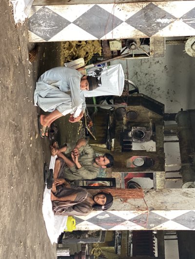 In a building in Shurandam, Afghanistan, three young men are resting next to an industrial machine. The floor and lower wall section feature a checkered black and white pattern. The first man is squatting with his arms around his knees, wearing a light-colored shalwar kameez and sandals, with a dark cap on his head. The second man is sitting cross-legged, wearing a similar shalwar kameez and holding his feet. The third man is also sitting cross-legged on a white sheet, wearing darker-colored clothes and a head covering. A yellow plastic container rests beside him. The industrial machine beside them appears to be in disrepair. A white sack leans against the first man.