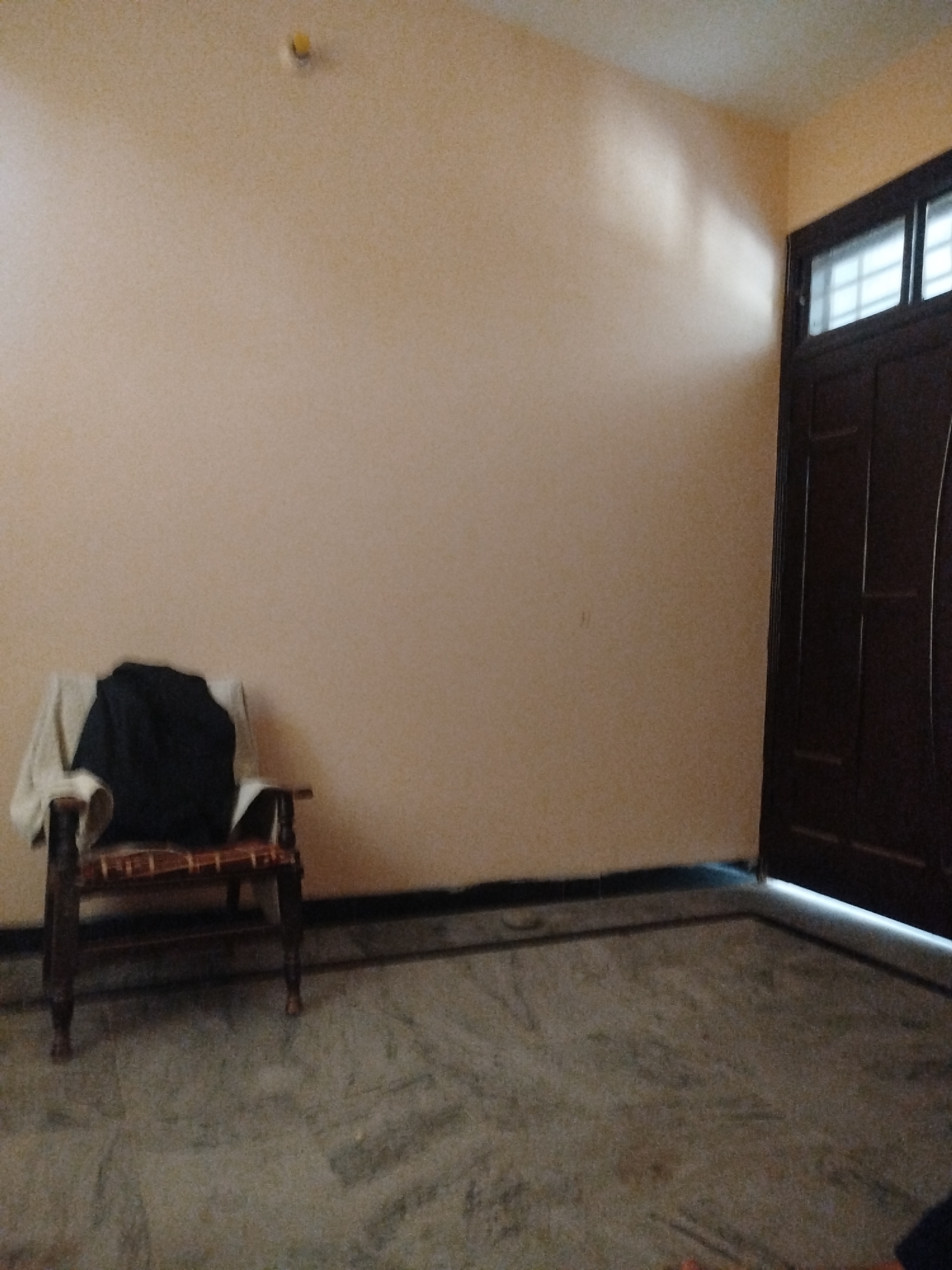 This is a full, eye-level, medium shot of an empty room. The walls are painted a light peach color. In the left half of the image, a wooden chair with woven seat and back is visible. A dark blue garment and a tan blanket are draped over the chair. The floor is tiled with large, grey, marble-like tiles. A dark brown double door with frosted glass windows is situated on the right side of the image. The scene appears to be a residential interior in Lassan, Pakistan, possibly a living room or bedroom.