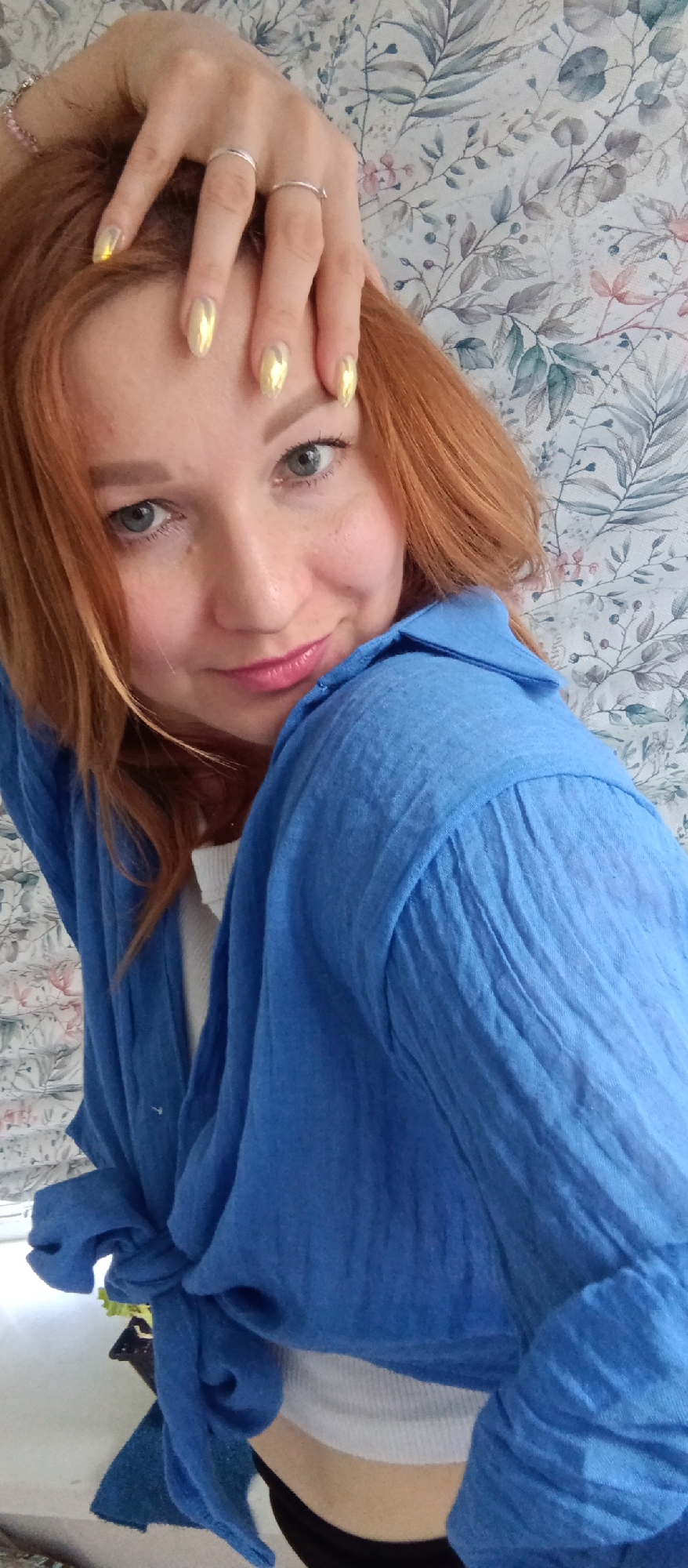 A close-up shot captures a woman with red hair and fair skin, posing indoors. She is wearing a loose-fitting blue shirt, partially tied at the waist, revealing a glimpse of a white undershirt underneath. Her left hand is raised, with fingers touching her forehead; two simple silver rings are visible on her hand. Her nails are painted with a gold or iridescent polish. Her face is angled towards the camera, displaying green eyes and light pink lipstick. The background features a wall covered in a floral pattern of green leaves, white blossoms, and muted pink hues. The city and specific location are not evident.