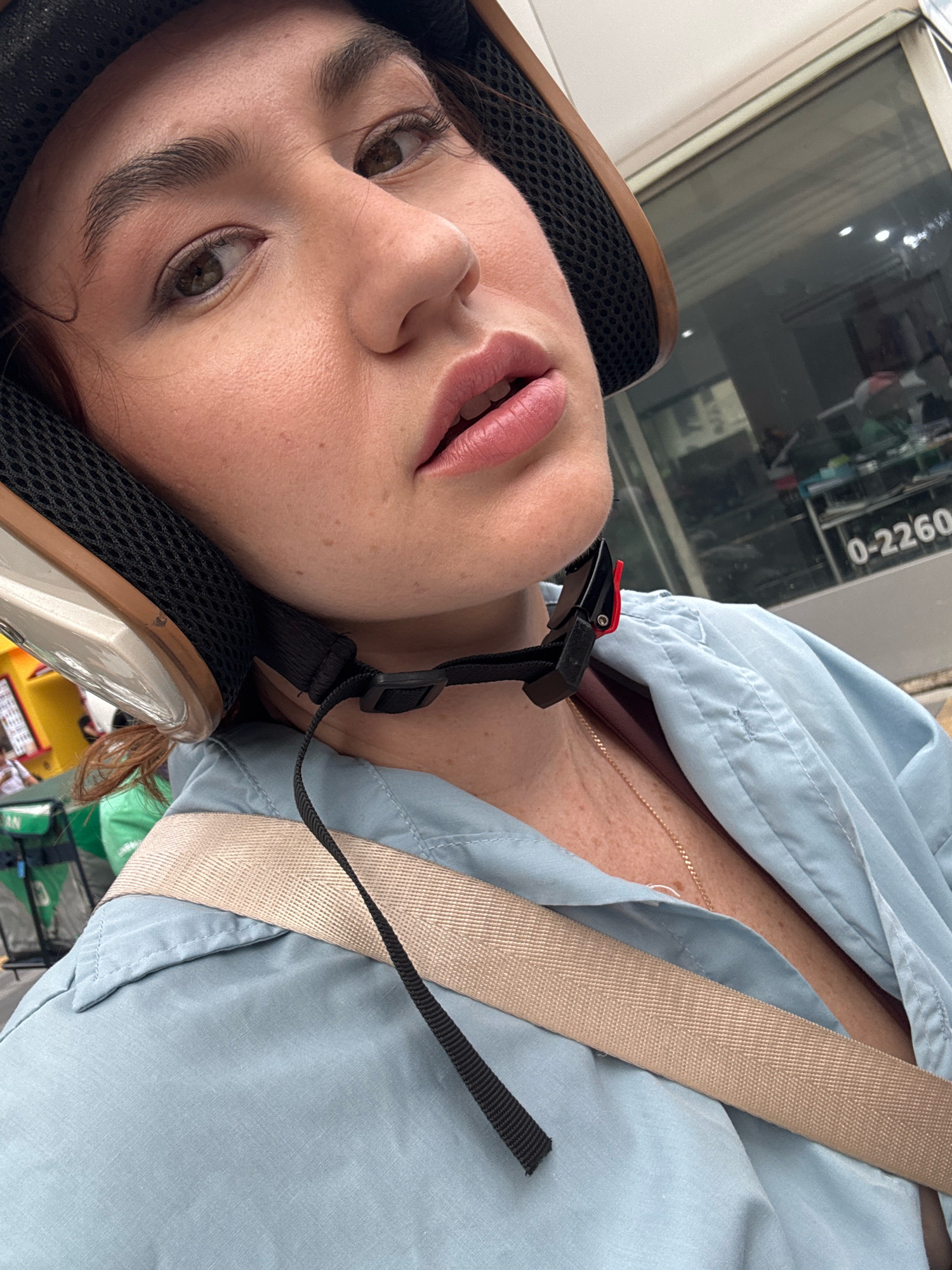A close-up, eye-level selfie shows a person wearing a motorcycle helmet. The person's face is angled towards the camera, with their lips slightly parted and their eyes looking away. Their skin is fair, with visible pores and freckles. They are wearing a light blue collared shirt with a tan strap across their chest. A gold necklace is visible around their neck. The background is blurred, but appears to be an outdoor urban environment. A glass building with a sign displaying the numbers "0-2260" is visible on the right. The scene is consistent with a location in Bangkok, Thailand, likely on a street or in a market area.