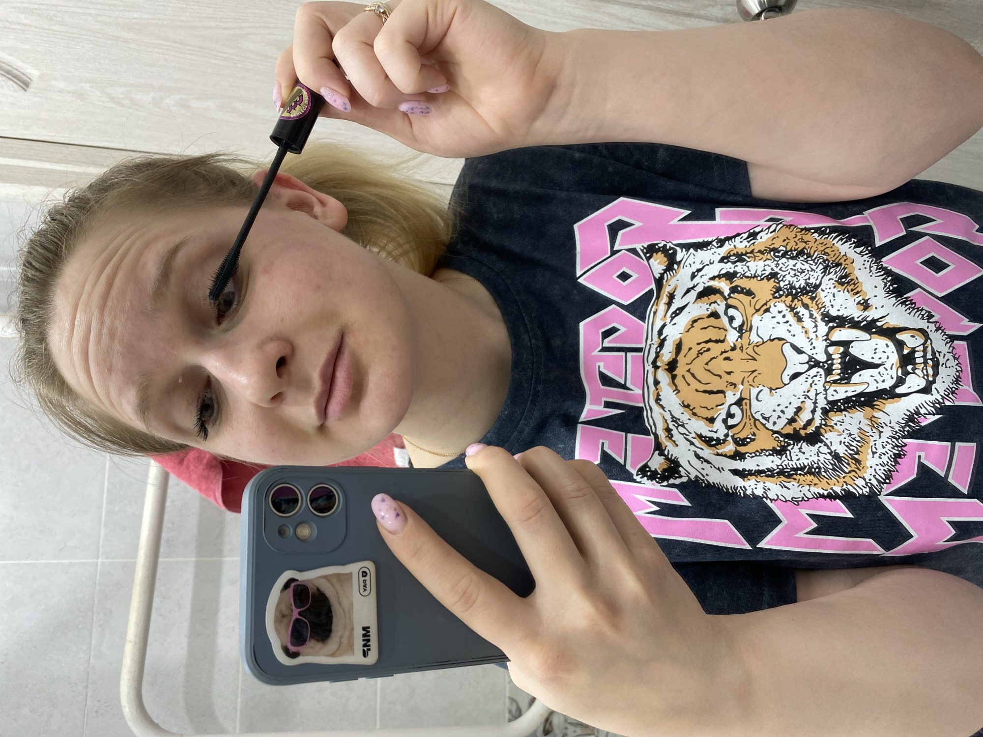 A young, light-skinned woman with blonde hair pulled back is applying mascara with her right hand. She is holding a grey smartphone in her left hand, presumably taking a selfie. The phone has a sticker of a pug wearing pink sunglasses on the back. She is wearing a dark grey t-shirt with a graphic print featuring a tiger and pink geometric shapes. The background appears to be a bathroom, with white tiles and a towel warmer visible. The location is Chaikovsky, Russia.