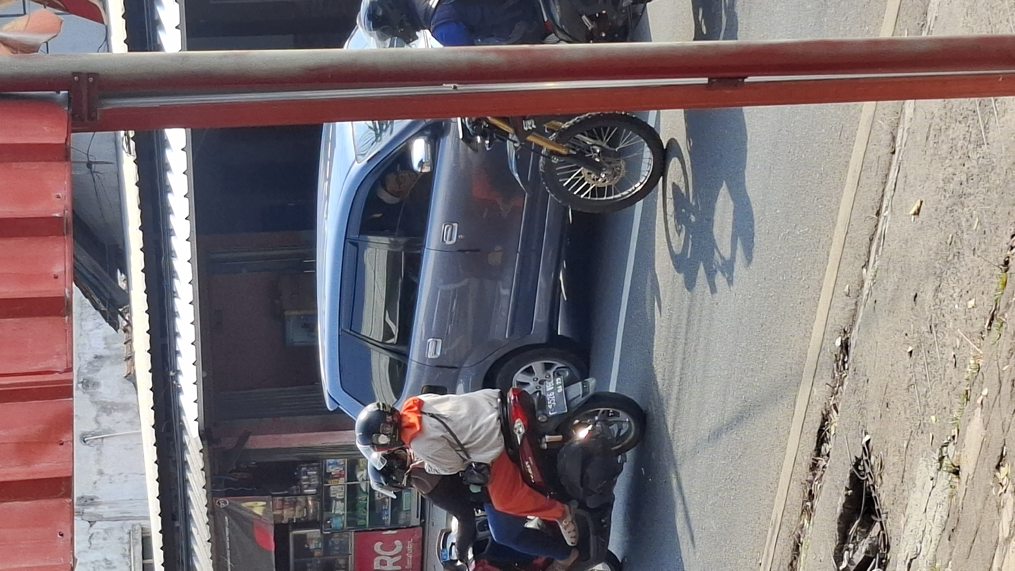 The image captures a street scene in Cilaku, Indonesia, on a bright, sunny day. In the foreground, a red metal pole with a red overhang is visible on the right side. The street is paved with asphalt, and a cracked section of the sidewalk with debris is present on the left.
The central focus of the image is a gray minivan stopped in traffic. Two individuals are riding a red scooter alongside the minivan. The passenger, a woman wearing a white jacket, orange headscarf, and orange pants, is looking towards the left. The driver, also wearing a helmet and dark clothing, is facing forward. Their scooter has a license plate that reads "F 5526 WBO".
To the right of the minivan, a dirt bike with yellow forks is stopped, its front wheel turned slightly to the left. The driver of this dirt bike is wearing a blue vest and a black helmet, looking towards the right. The background shows buildings with corrugated metal roofs and storefronts, some displaying merchandise. The overall atmosphere suggests a typical day on a busy Indonesian street.