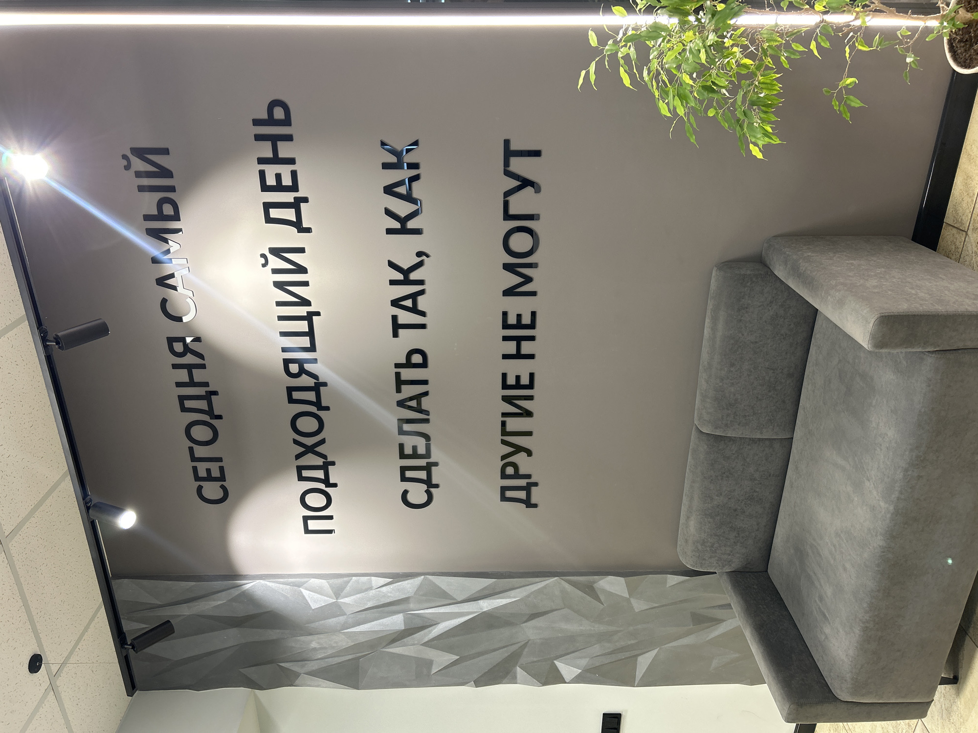 A photograph taken indoors at a location in Votkinsk, Russia, shows a waiting area with a gray sofa, a wall with raised geometric patterns, and a plant. The text on the wall reads "СЕГОДНЯ САМЫЙ ПОДХОДЯЩИЙ ДЕНЬ СДЕЛАТЬ ТАК, КАК ДРУГИЕ НЕ МОГУТ". The lighting comes from track lighting on the ceiling.