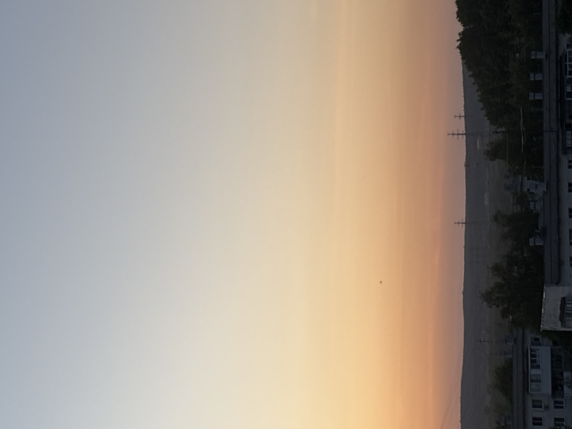 The image shows an early morning or late evening sky over Chaikovsky, Russia. The sky fades from light blue to orange near the horizon. A treeline and buildings are visible in the lower right portion of the image, along with power lines in the background. The time of day appears to be sunrise or sunset due to the sky's color.