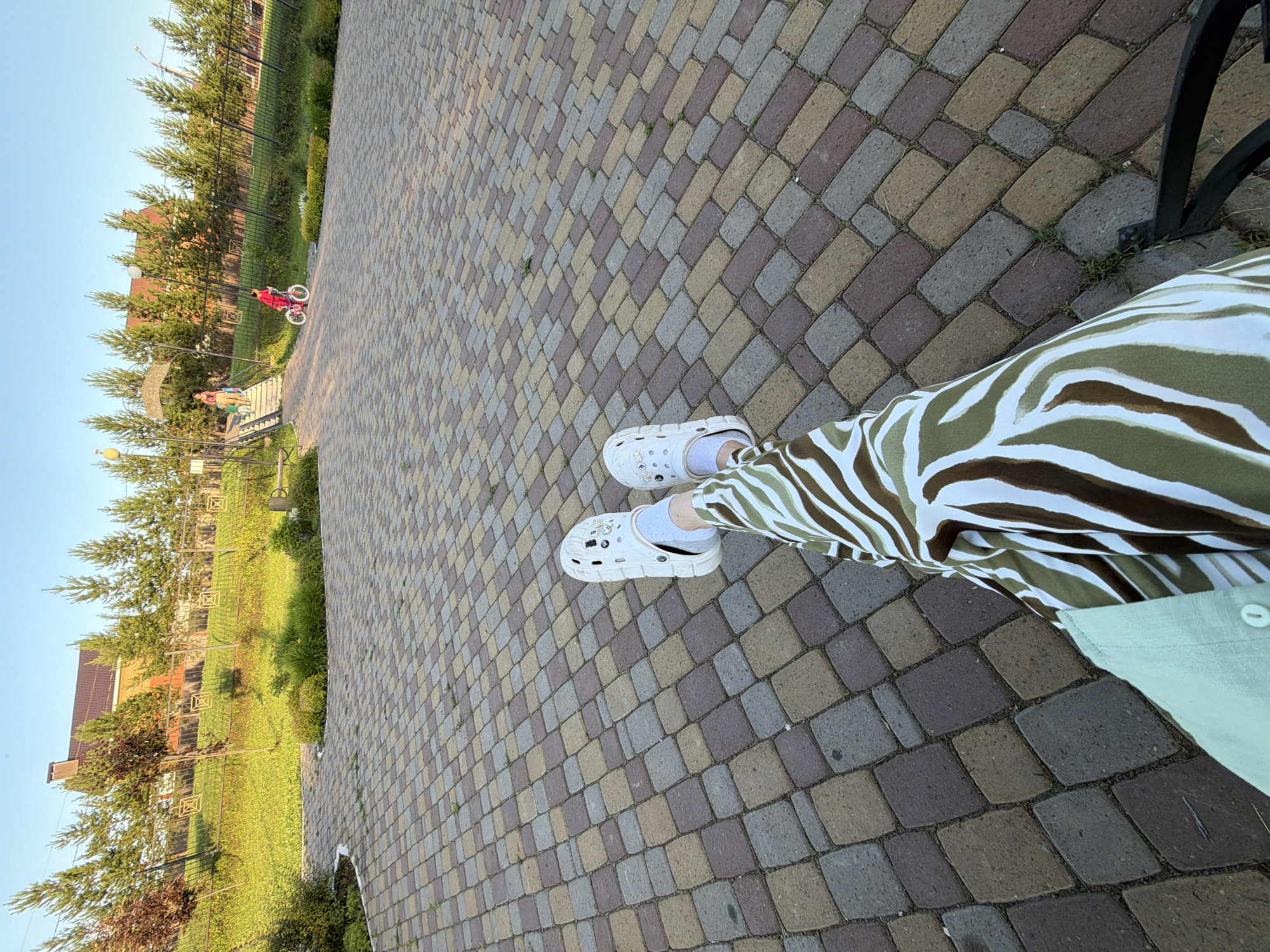 An outdoor, eye-level shot shows a person's legs and feet sitting on a bench in a park. The person is wearing white Crocs and green and white zebra-patterned pants. The park has a paved walkway with multicolored cobblestones. In the background, a fence runs along a grassy hill, with trees and a house visible beyond. A small child on a red bicycle rides on the walkway in the distance, and another child and adult are walking up a set of stairs. The sky is clear and blue.