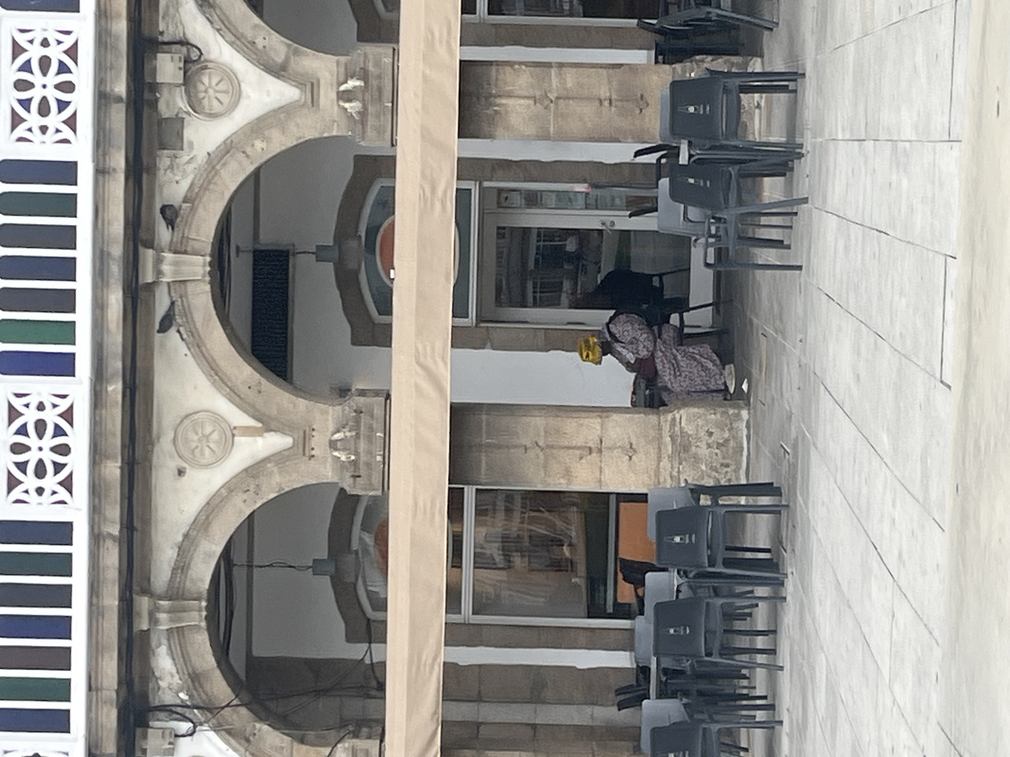 The image shows a section of the arcaded gallery in María Pita Square, A Coruña, Spain. The arches are made of granite, with decorative rosettes. There are several chairs stacked nearby and a person seated in front of a window in the background. The building’s facade features a modern awning and stained glass elements.