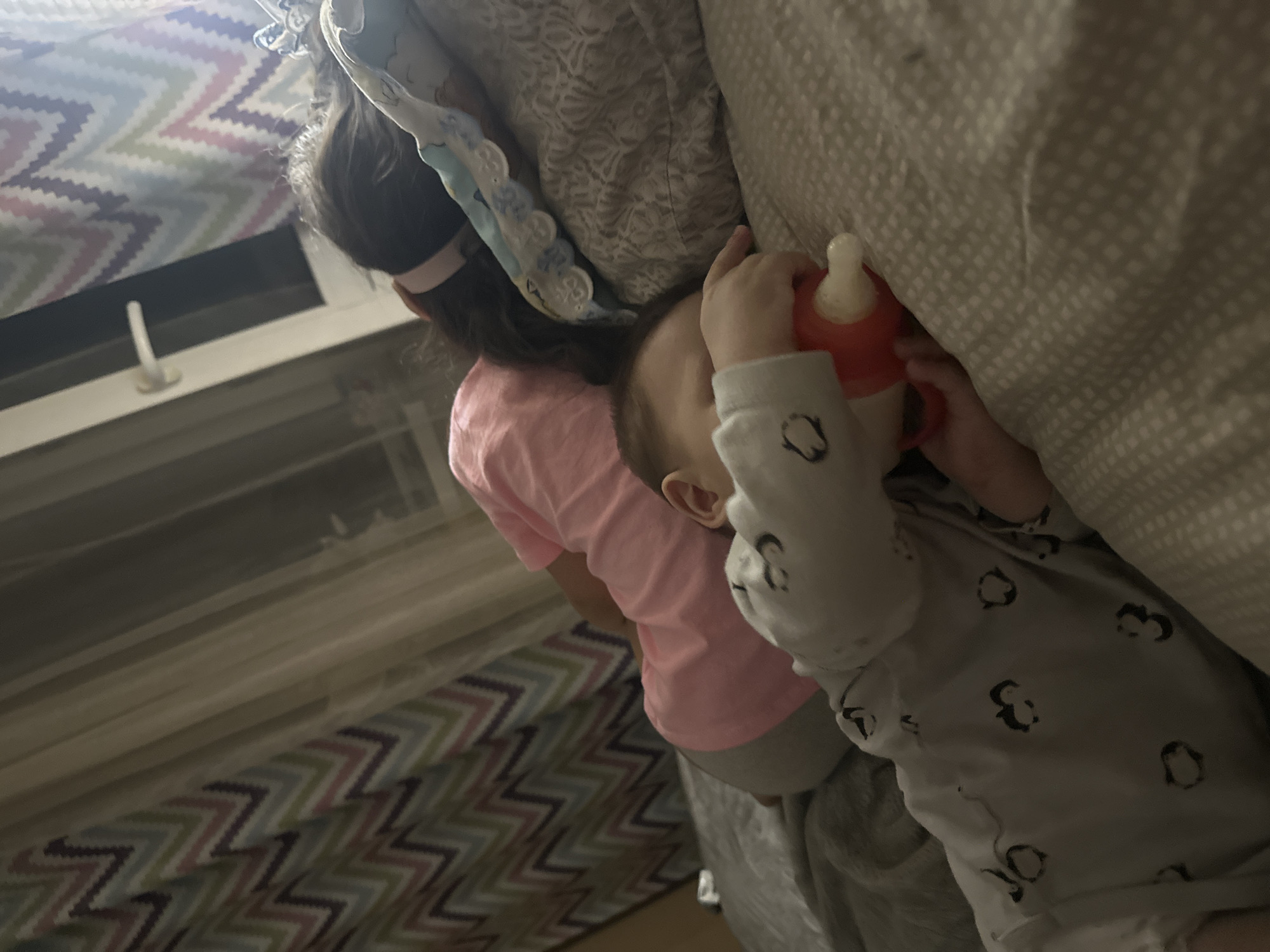 The image depicts a child in penguin-printed pajamas lying on a bed with a patterned gray and white comforter, drinking from an orange bottle. Another child wearing a pink shirt stands near the first child. The background includes a window and a patterned curtain. The location is unknown.