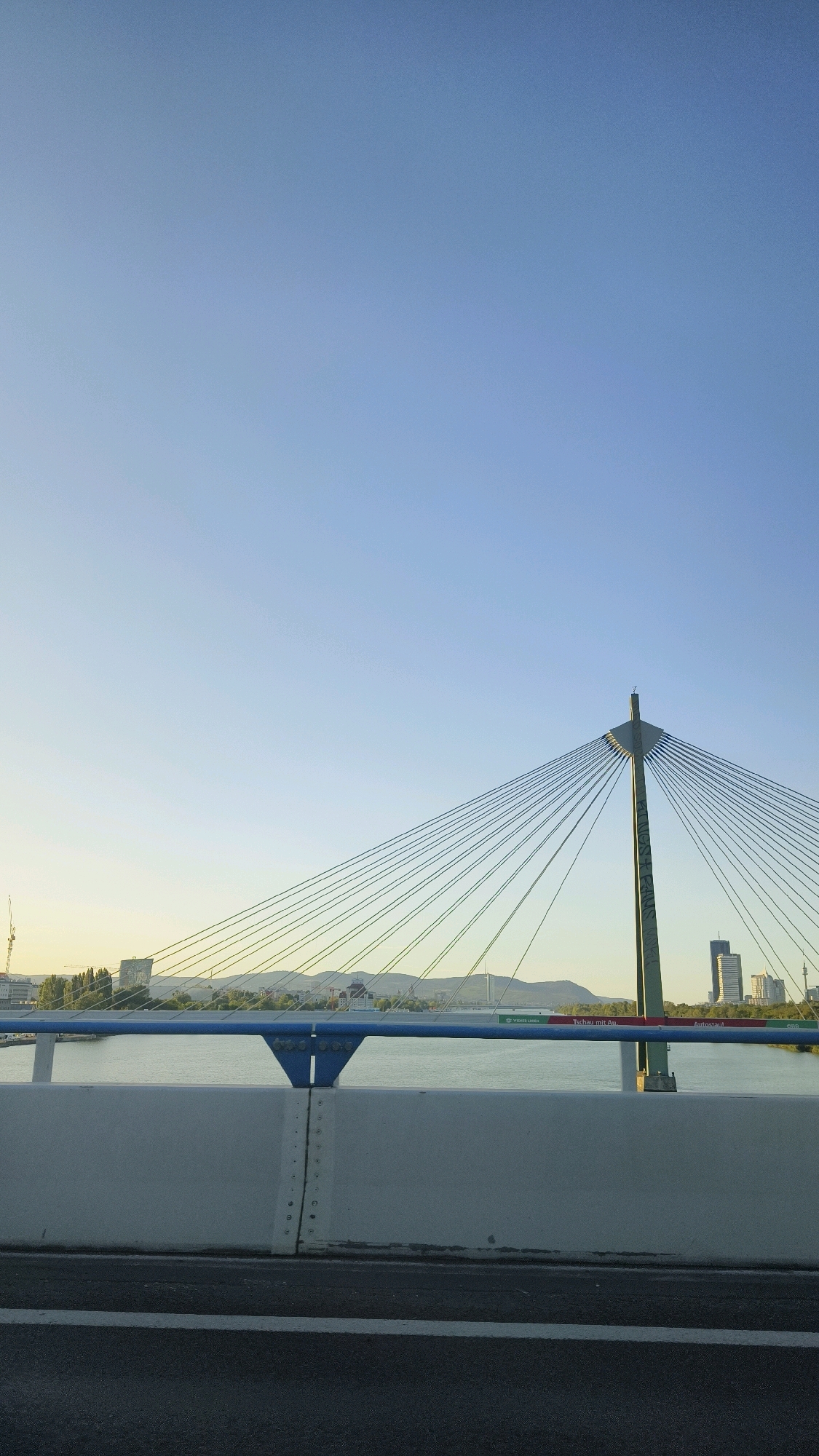 This is a vertical, eye-level, outdoor, daytime photograph taken from a bridge. The sky is a clear, light blue with a subtle gradient. In the middle ground, a suspension bridge's central pylon rises, with numerous cables fanning out from its top and anchoring points. The pylon is a tall, dark green structure with some graffiti visible on its surface. Below the bridge cables, a body of water, likely the Danube River, is visible. Along the far bank of the river, a city skyline features trees, several buildings, including two prominent high-rise towers, and distant hills. The foreground of the image is dominated by the bridge deck the viewer is presumably on, showing a grey concrete barrier with blue railings and a section of asphalt road with a white stripe. The overall scene suggests a view of Vienna, Austria, encompassing its urban landscape and the Danube River. There are no people or animals visible in the image.