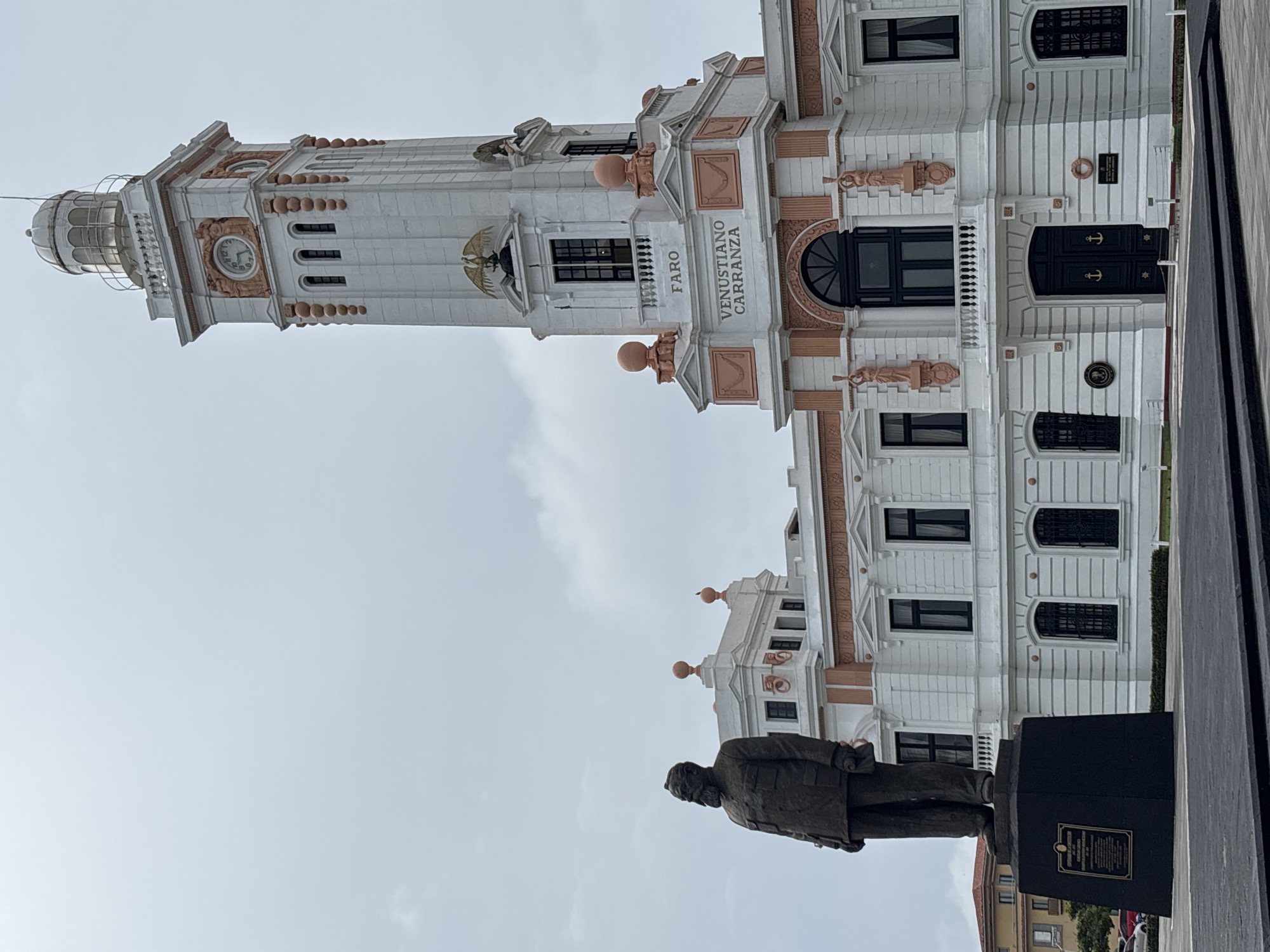 This is a photograph of the Faro Carranza, a lighthouse and administrative building, in Veracruz City, Mexico. The image is oriented vertically, with the sky taking up the upper portion and the building and a statue occupying the lower portion. The Faro Carranza is a tall, white, ornate structure with pink accents and a dome-shaped lantern room at its peak. The building features multiple windows, balconies, and decorative elements, including statues and what appear to be eagles. The words "VENUSTIANO CARRANZA FARO" are visible on the side of the building. In the foreground, a bronze statue of a man in a suit stands on a dark pedestal. The statue faces to the left, away from the camera. The sky is overcast, suggesting a cloudy day. The scene appears to be a public space, likely a plaza or waterfront area, given the presence of the monument and the building's grandeur. The time of day is not clearly discernible due to the overcast conditions, but it appears to be daytime.