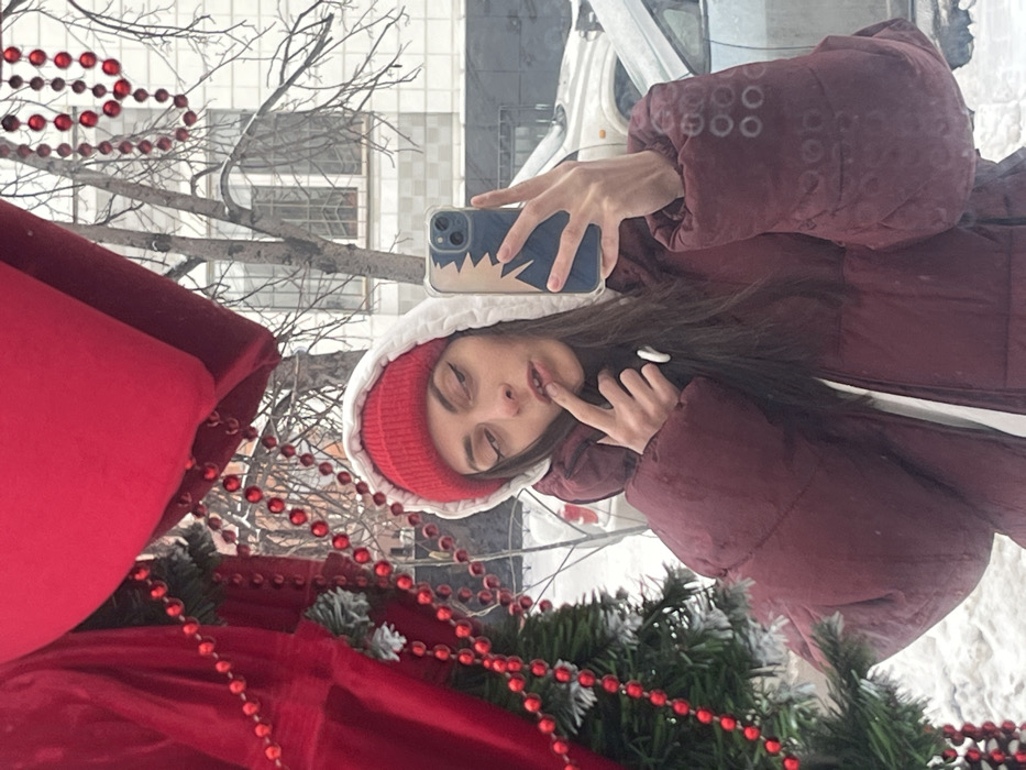 A young woman in a red beanie and maroon puffer jacket takes a selfie. She’s posing with a finger to her lips. Her phone has a blue and white case. The reflection shows a winter scene with bare trees and a building. A red fabric and green garland with red beads are visible, possibly holiday decorations. The overall image has a slightly tilted, selfie perspective.