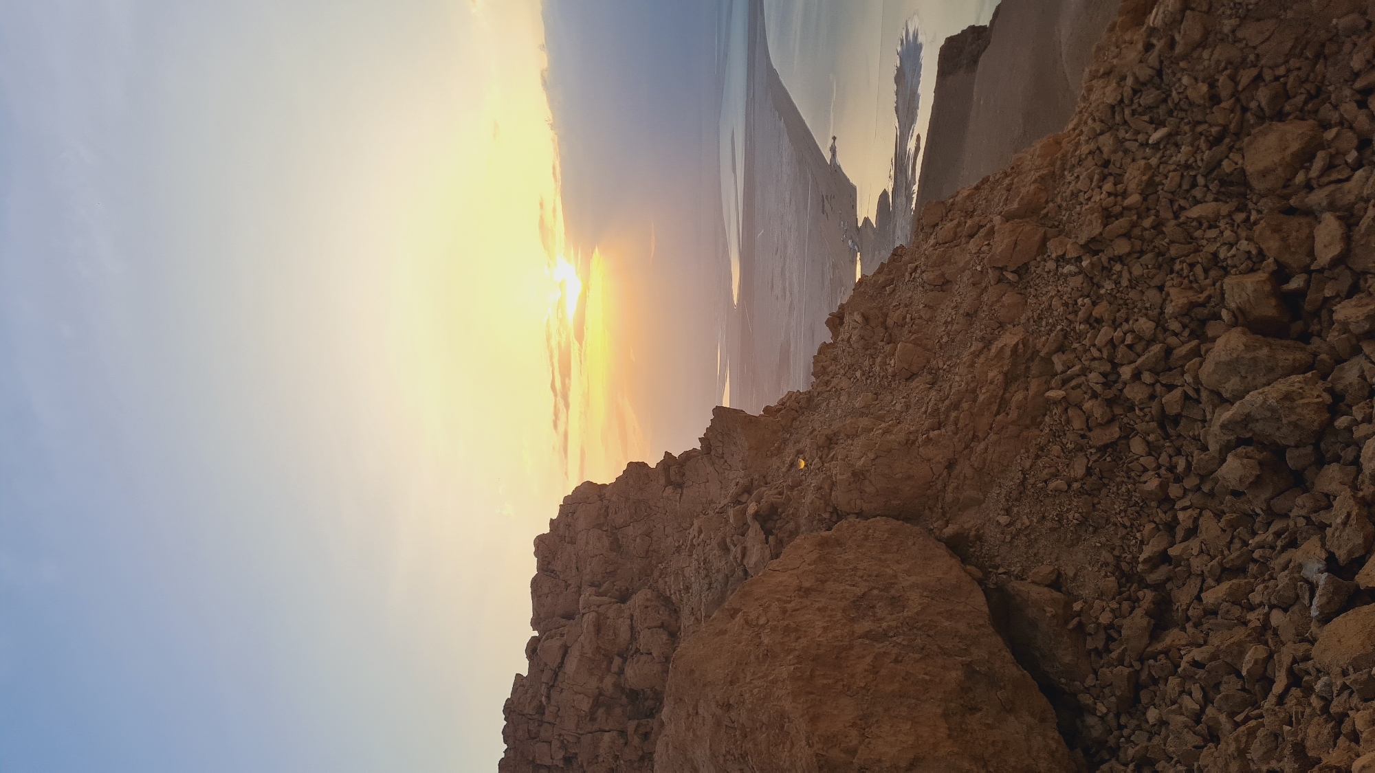 The image shows a rocky landscape at what appears to be the edge of a cliff or elevated terrain. The foreground consists of various sizes of rocks and stones, in shades of brown and tan. In the background, a body of water stretches into the horizon, reflecting the sunlight. The sky transitions from a light blue to a yellow-orange hue, indicating either sunrise or sunset. The sun itself is partially obscured by clouds, creating a bright glare in the sky. Given the location is Tamar Regional Council, Israel, the body of water may be the Dead Sea. No people, interactions, or other discernible subjects are visible.