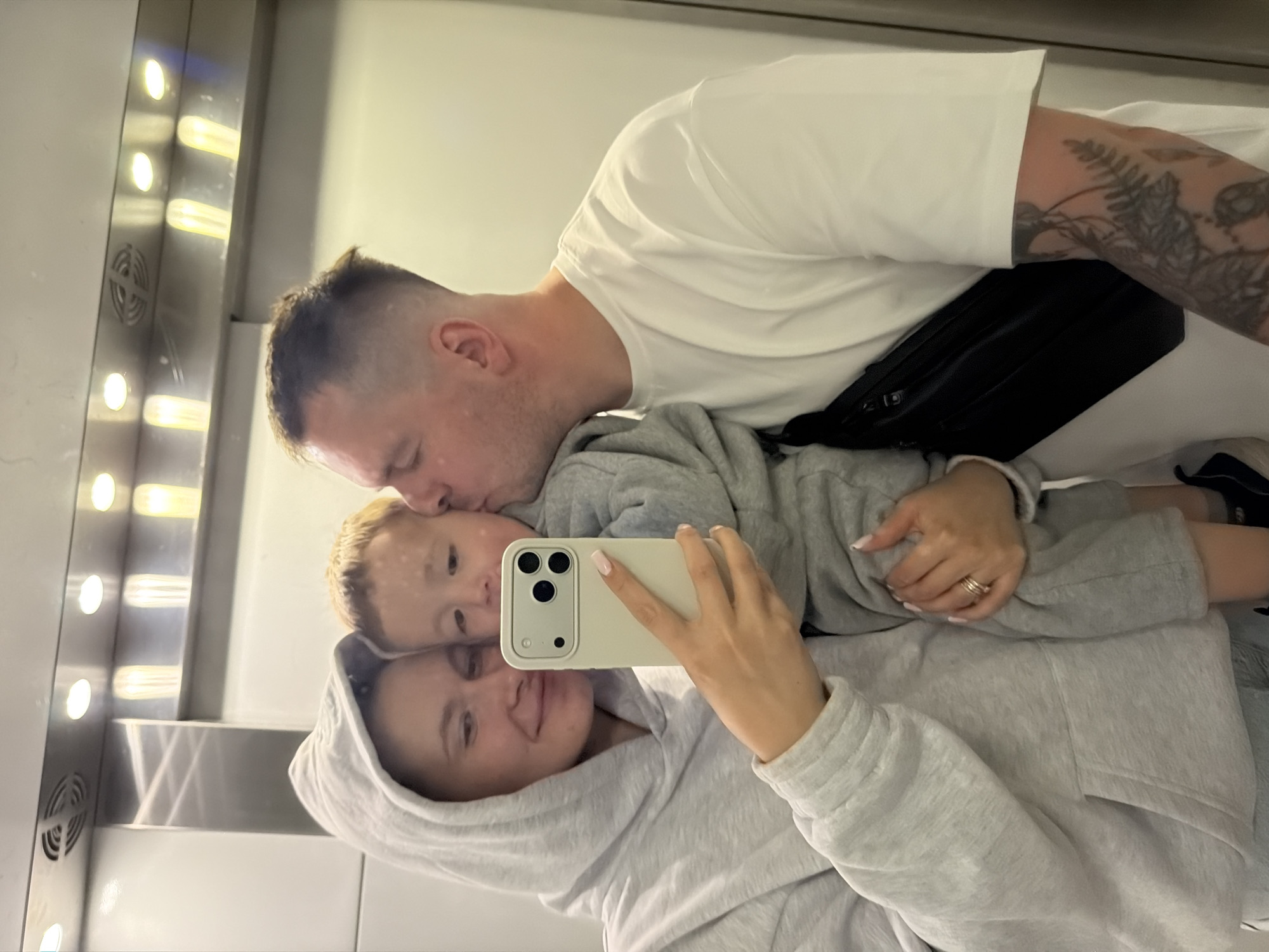 This image, taken in Odintsovo, Russia, captures three individuals reflected in a mirrored surface, likely a selfie. The scene depicts an adult female, a young child, and an adult male.
The adult female, positioned on the left, wears a light grey hooded sweatshirt with the hood up. She has light-colored hair, visible underneath the hood. She is holding a light-beige smartphone with a triple camera array in her right hand, seemingly taking the photograph. Her fingernails are manicured with a light pink polish, and she wears a triple-band ring on her left ring finger. She exhibits a faint smile and gazes towards the phone.
The young child, a toddler with light-colored hair, is held between the female and male. The child also wears a light grey hooded sweatshirt and looks towards the phone.
The adult male, positioned on the right, has short, dark hair with faded sides. He is observed kissing the child's right cheek. He wears a white short-sleeved t-shirt and a black fanny pack or sling bag across his chest. Tattoos, depicting botanical or vine-like patterns, are visible on his right forearm.
The background, visible through the reflection, consists of light-colored walls. Above the subjects, a metallic panel features several recessed circular lights emitting white light, interspersed with vertical linear light fixtures and a circular ventilation grate. This arrangement suggests an interior space, possibly an elevator or a hallway with reflective architectural elements.