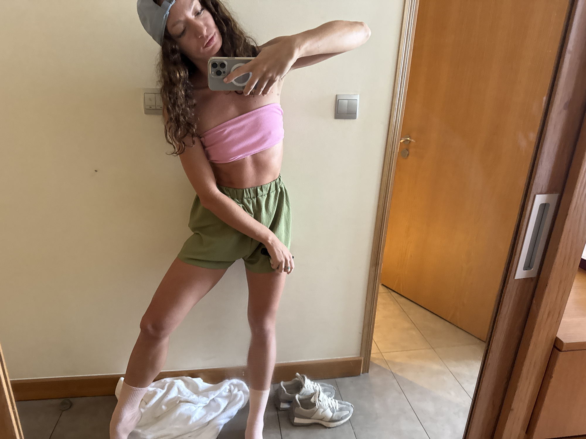 A young woman takes a selfie in a mirror. She wears a light blue baseball cap, a pink tube top, and green shorts. She stands in socks on a tiled floor. Behind her are a pair of gray and white sneakers and a rumpled white cloth. A light switch is visible on the wall. A wooden door is partially open. The scene is likely a room in an apartment or hotel. Đà Nẵng, Vietnam is not discernible from the image.