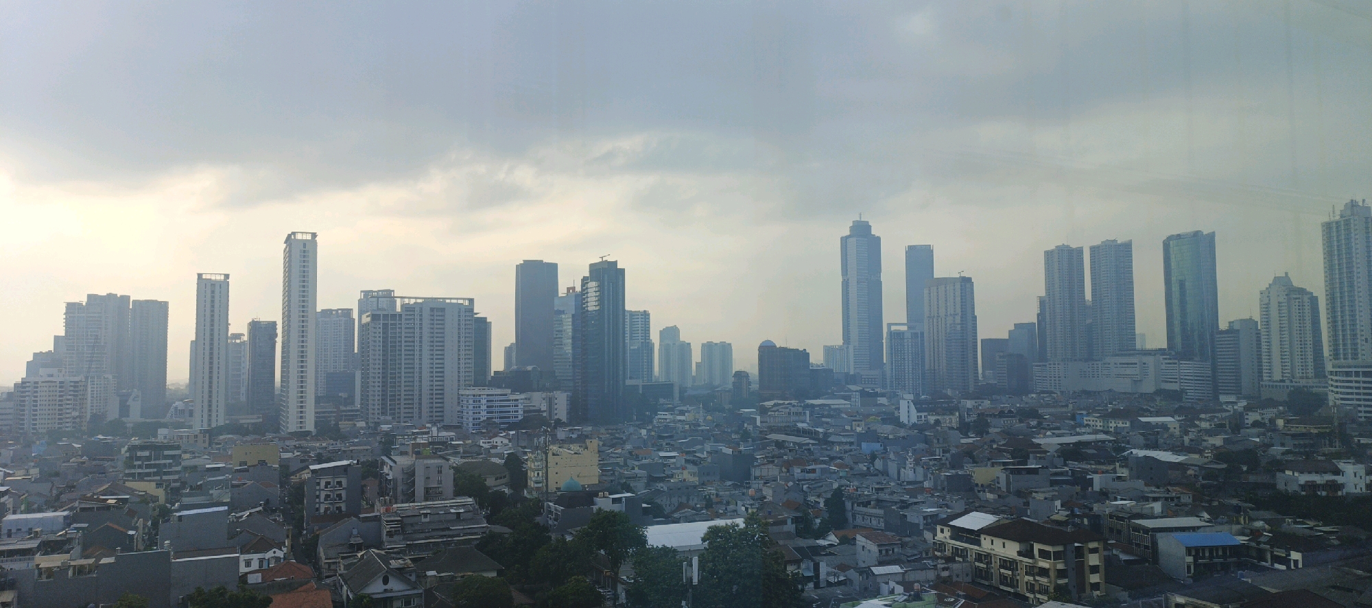 This is a panoramic, high-angle shot of the Jakarta skyline, likely taken from a tall building, overlooking a dense urban area of South Jakarta, Indonesia, under a hazy, overcast sky. In the foreground, a dense cluster of low-rise buildings, with varying roof colors and architectural styles, fills the bottom half of the frame. These structures are predominantly made of concrete and appear to be residential or commercial.
Behind the low-rise buildings, a dramatic contrast emerges as a collection of modern skyscrapers rises into the sky. These high-rise buildings are of diverse designs, some with sleek, glass facades reflecting the muted light, while others are more utilitarian in appearance with visible structural elements. The skyscrapers vary in height and form, creating a complex and layered cityscape.
The environment is characterized by a lack of distinct weather phenomena, but the haze suggests a potential for pollution or morning fog, obscuring distant details and lending a soft, diffused quality to the light. No individuals are clearly visible in the frame, but the presence of numerous buildings implies a populated urban environment. The overall impression is one of a bustling, developing metropolis, where older neighborhoods coexist with modern high-rise development.