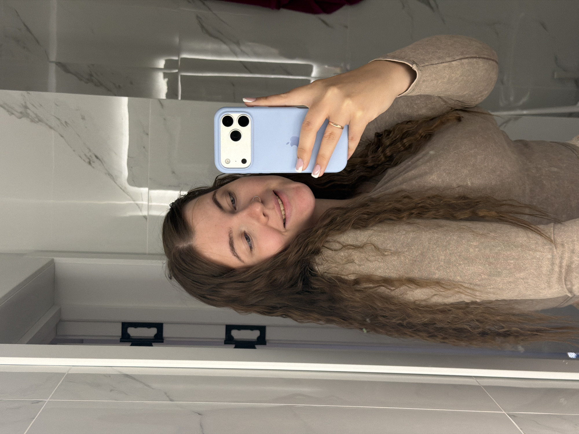 A woman with long, curly brown hair is taking a selfie in a bathroom with gray and white marble-patterned tiles. She is holding a light blue iPhone with a triple-lens camera. She is wearing a beige shirt and a ring on her ring finger. The location is possibly in Chaikovsky, Russia.