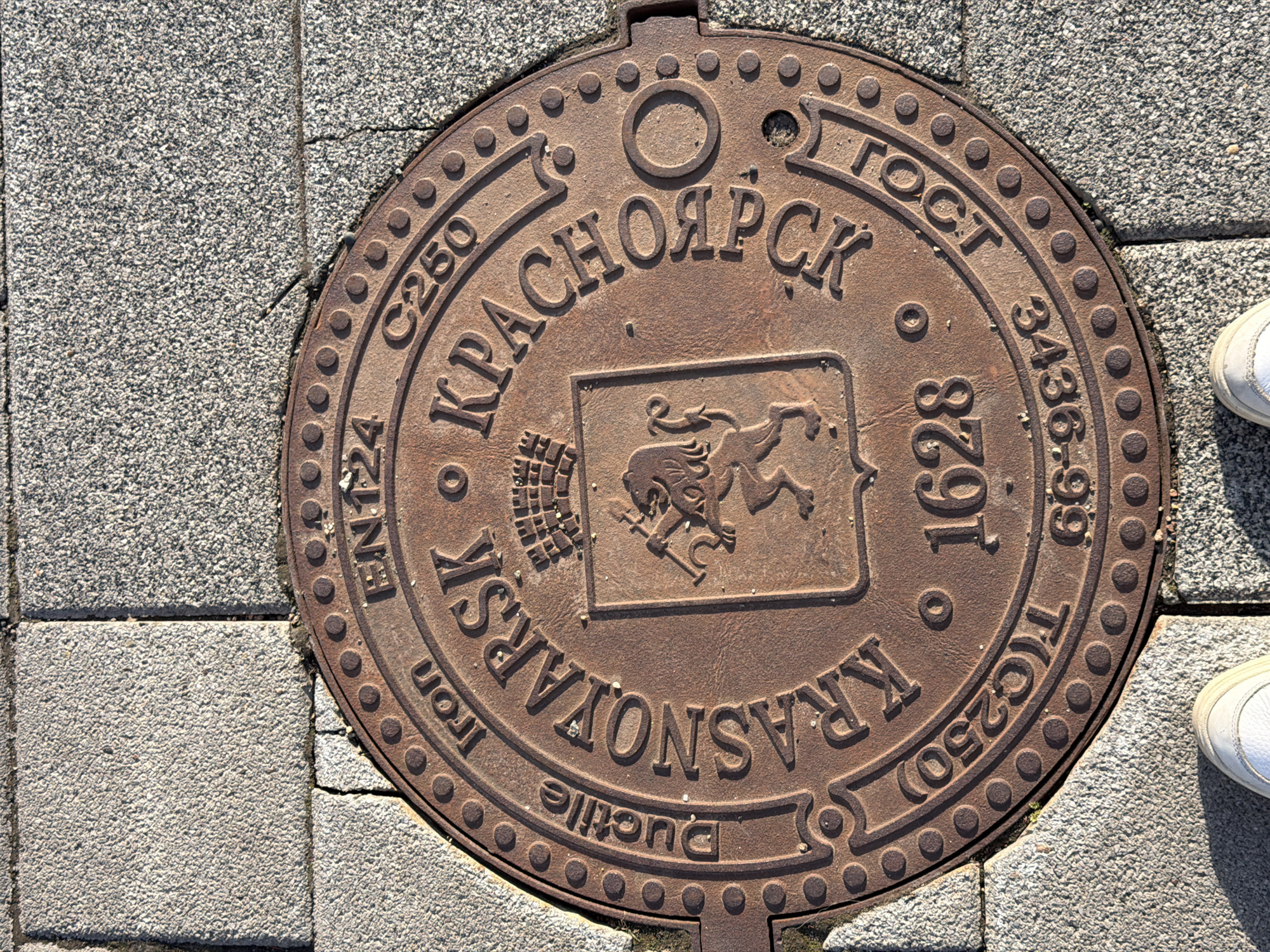 A top-down shot depicts a round, rusty-brown manhole cover set into a gray, interlocking brick walkway. The cover prominently displays raised lettering spelling out "КРАСНОЯРСК" in Cyrillic, and "KRASNOYARSK" in Latin letters. A central square shield features a heraldic lion figure holding a shovel and sickle. Other lettering and numbers, including "EN124 C250", "ГОСТ 3436-99 T(C250)", and "1628" are embossed around the perimeter. A pair of white sneakers is visible to the right of the cover. The location is most likely Krasnoyarsk, Russia.