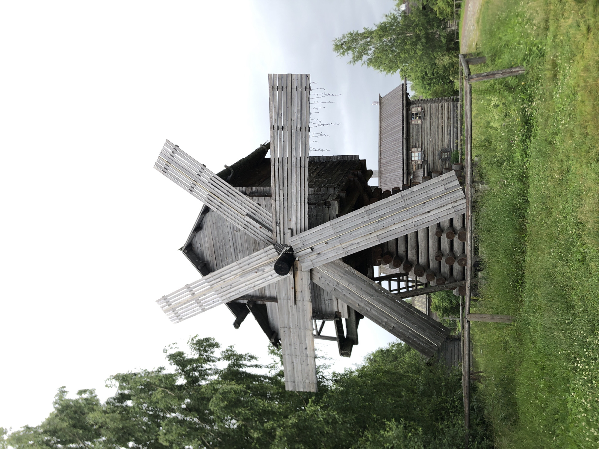 This is a photograph of an outdoor scene featuring a traditional wooden windmill and a log house. The windmill is the central subject, with its large wooden blades prominently displayed. The blades are constructed from planks of wood, creating a latticed appearance. The windmill appears to be made of weathered wood, with a dark, aged roof.
Adjacent to the windmill is a rustic log house. It has several small windows with visible panes and appears to be a historical structure, possibly part of an open-air museum or heritage village. The overall construction of both buildings is wooden, suggesting a rural or historical setting.
The surrounding environment consists of lush green grass and trees, indicating a natural, possibly rural, location. There are some small white flowers scattered in the grass. A wooden fence or railing is visible in the foreground, separating the grassy area from the structures.
The sky is overcast and appears pale, suggesting a cloudy day, possibly in the morning or late afternoon. There is no visible text in the image. The scene is static, with no people or animals actively engaged in any activity. The focus is on the architectural elements and the natural surroundings. The orientation of the image is landscape. The location context provided is Verkhniye Mandrogi, Russia.