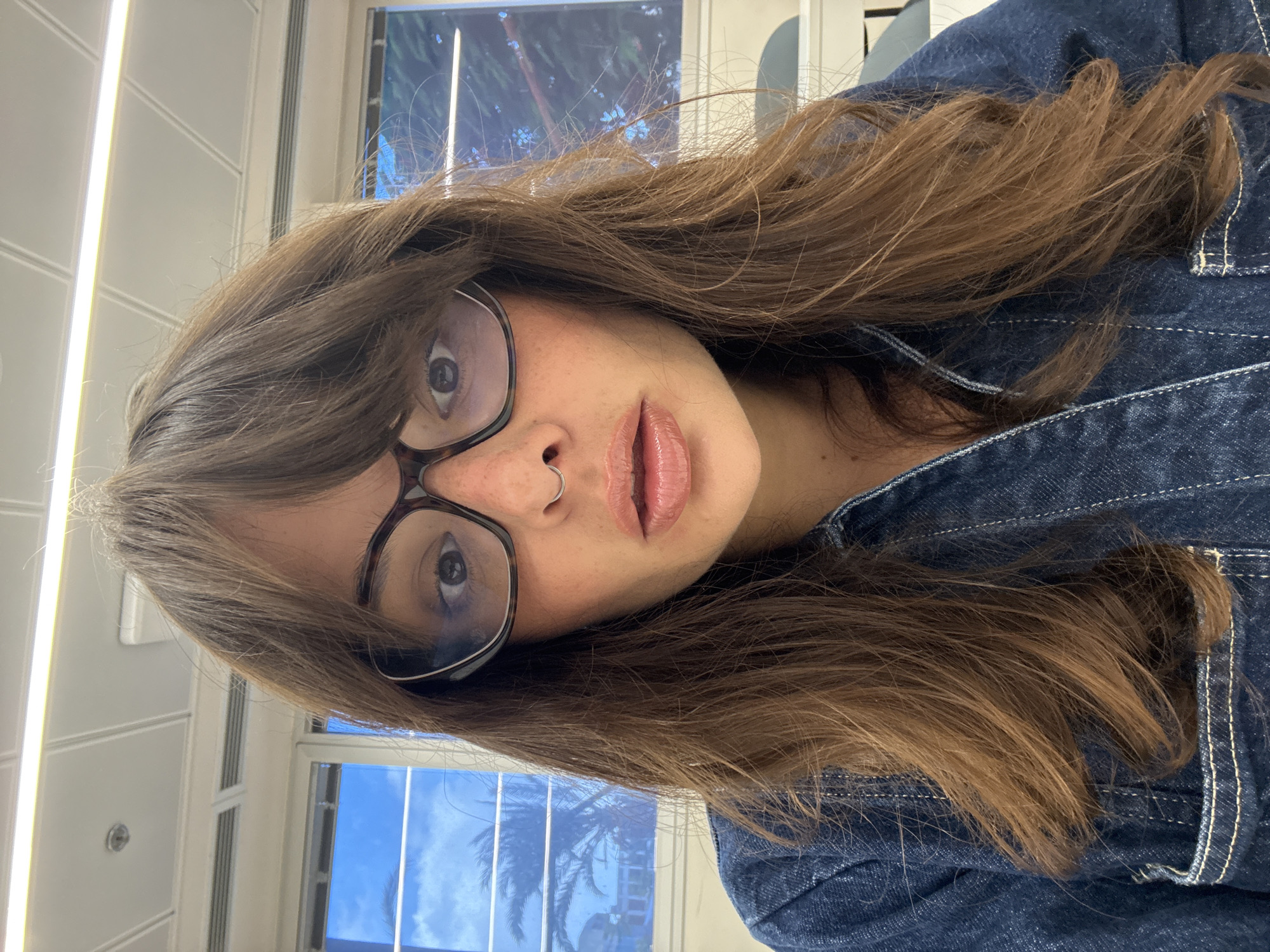 This is a close-up, head-on selfie of a young woman with brown hair, bangs, and glasses. She is wearing a denim shirt and has a nose ring. Her expression is neutral, and she is looking directly at the camera. The background appears to be an indoor setting with large windows. Through the windows, palm trees and a bright blue sky are visible, suggesting a warm, possibly coastal climate. The interior features fluorescent lighting and what appear to be office or classroom elements like partition walls. The overall impression is of a casual, everyday moment captured in an urban environment. Given the presence of palm trees against a clear blue sky, this scene could be located in a modern, sunny city. Tel Aviv, with its prominent coastline and modern architecture, fits this description well.