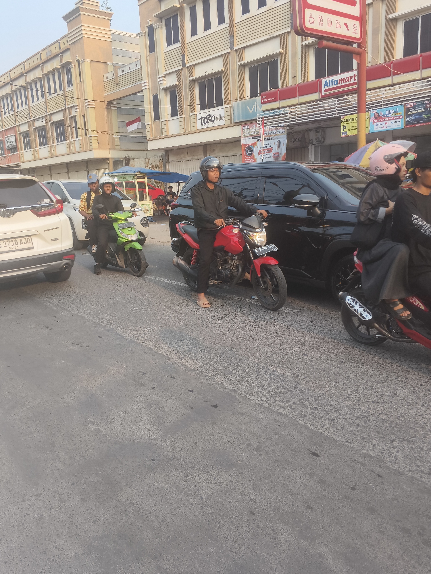 A street scene in Bandar Lampung, Indonesia, shows several vehicles and people in traffic. In the foreground, a person on a red motorcycle, identified by license plate B 3235 VHN, is moving with a black SUV to their right. To the left of the red motorcycle, a green scooter carrying two people, one wearing a yellow shirt, is stopped. Further to the left, the rear of a white SUV, license plate BE 1718 AJO, is visible. In the background, buildings with commercial signage, including "Alfmart" and "TORO GALLERY," line the street. A small cart with a blue canopy is also present. The sky is overcast.