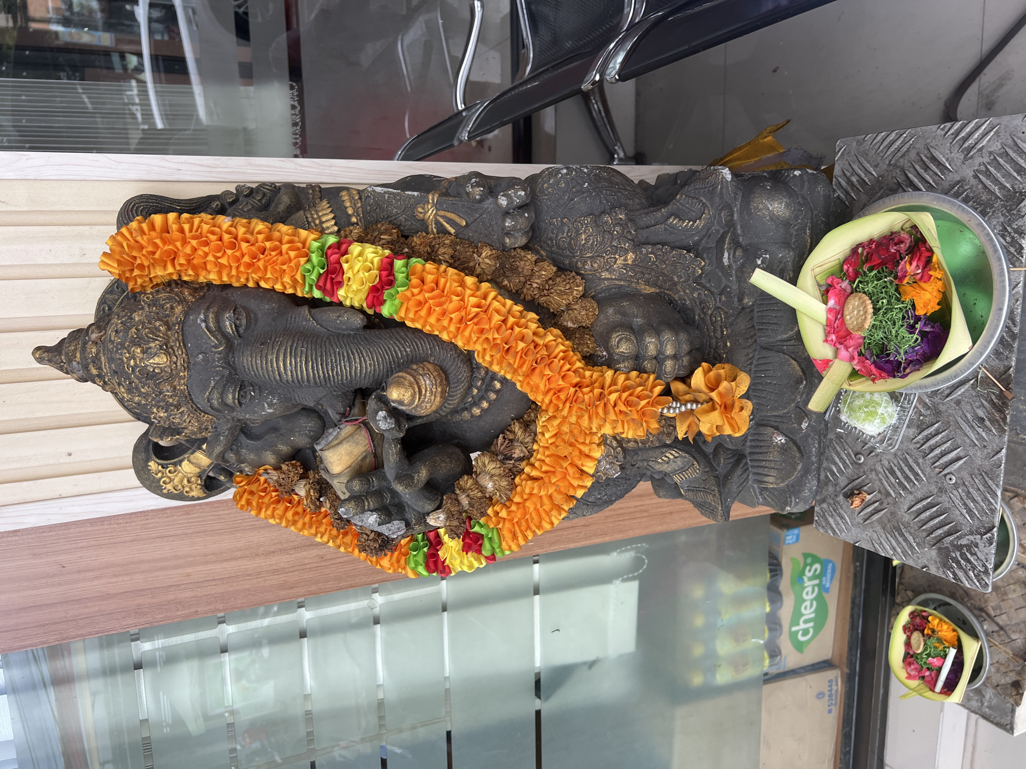 A dark, intricately carved statue of Ganesha, adorned with colorful garlands, is the central focus of the image. The statue, possibly made of stone or concrete, features golden accents on its trunk, crown, and ornaments. One prominent garland is bright orange, made of ruffled fabric, encircling Ganesha's neck and draped over his body. Another garland visible has segments of red, yellow, and green fabric, and a third appears to be made of dried floral or plant material. Ganesha is depicted in a reclining or seated pose, with offerings placed beside him.
To the right of the statue, on a metallic, textured surface, are two traditional Balinese Hindu offerings known as Canang Sari. These are small, round baskets woven from palm leaves, each filled with a vibrant assortment of fresh flowers (including red, pink, orange, and purple petals), green foliage or herbs, and a small, round biscuit. One offering also contains a thin white stick, possibly incense. A small, light green, spherical dessert item in a clear plastic container is also present on the metallic surface.
The background shows reflections in glass panels, indicating an indoor or semi-outdoor setting. Reflected in the glass are black chairs with chrome legs, and a cardboard box with the word "Cheers" visible on its side. The statue rests on a wooden surface, suggesting a display area. The overall scene conveys a sense of reverence and cultural practice, consistent with offerings in Kuta Selatan, Indonesia.
