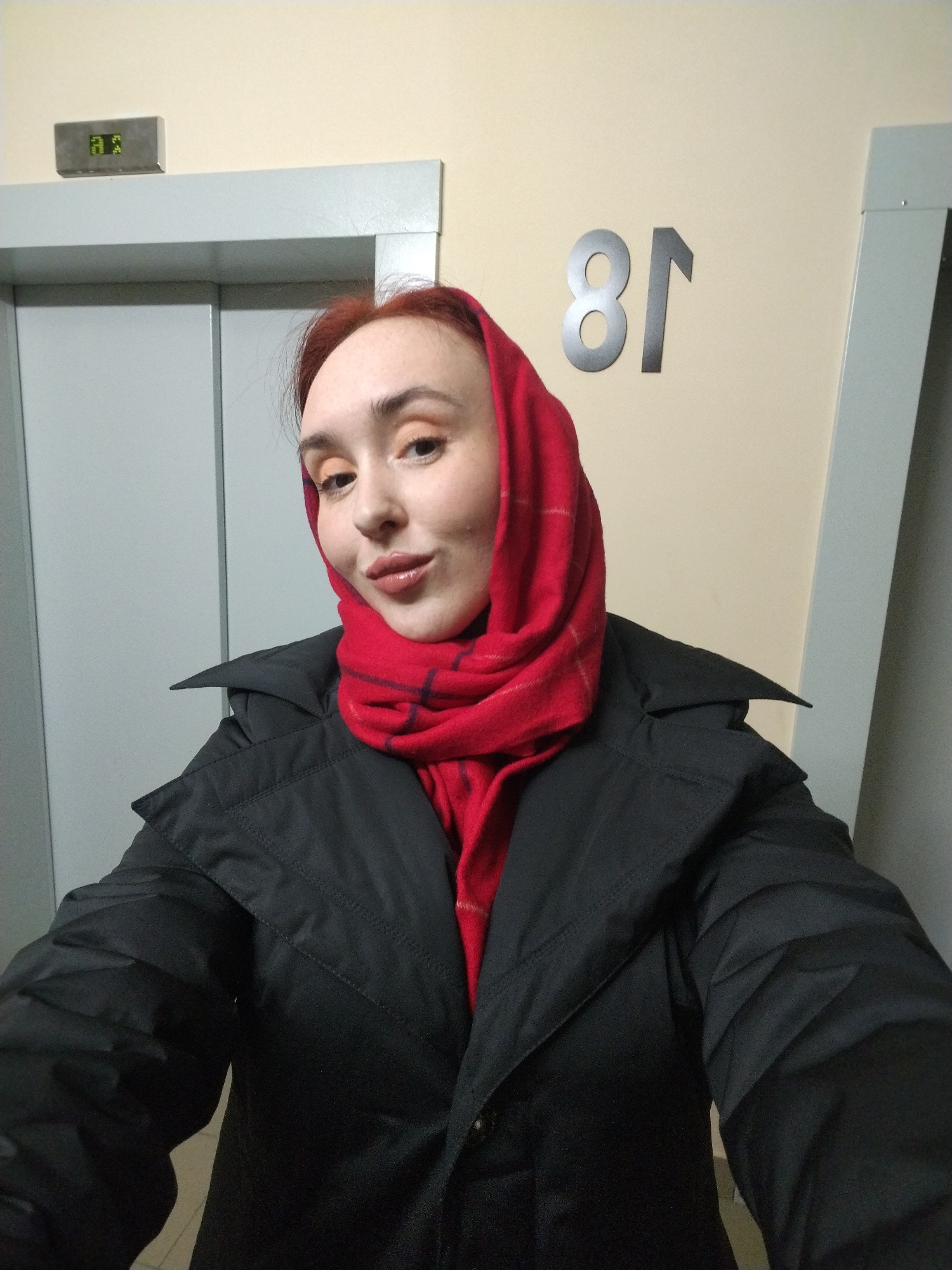 A woman wearing a black jacket and a red headscarf is standing in front of an elevator. The elevator is located in a building, with the number 81 visible on the wall. The building is located in the city of Izhevsk, Russia.