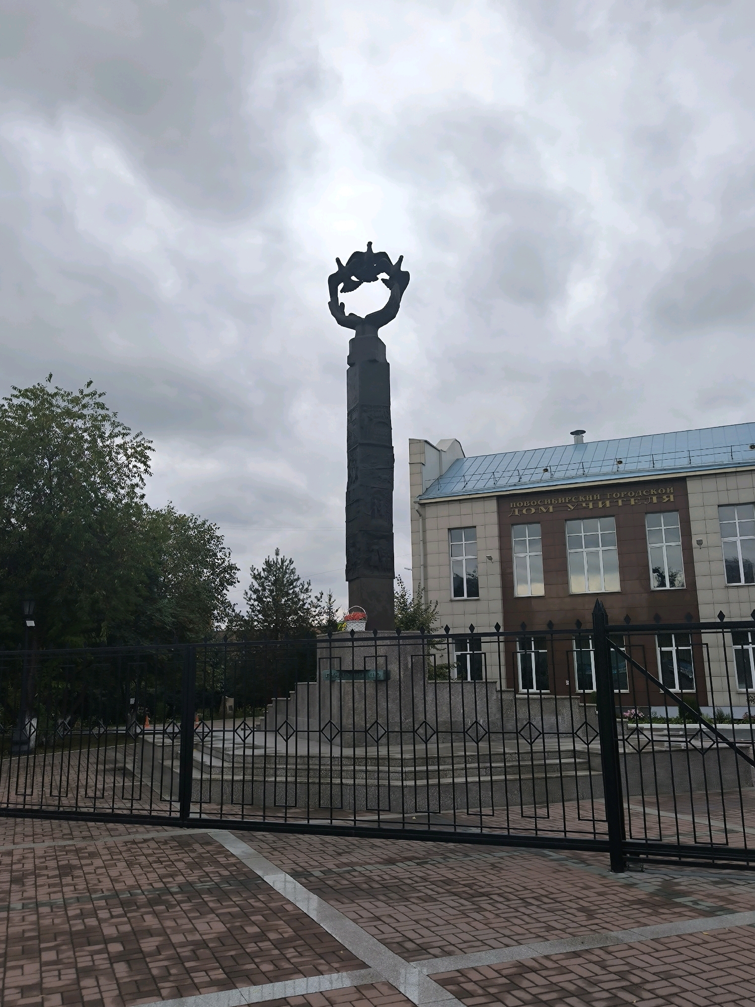 The image shows a view of a monument and a building in Novosibirsk, Russia. The monument is a tall, dark obelisk topped with a sculpture of two figures, possibly birds or winged beings, in a circular formation. The obelisk is adorned with relief carvings along its shaft. It stands on a tiered base. To the right of the monument is a light-colored building with a blue roof. The building has multiple windows and an inscription in Russian that reads "НОВОСИБИРСКИЙ ГОРОДСКОЙ ДОМ УЧИТЕЛЯ" (Novosibirsk City House of Teachers). A black metal fence runs across the foreground, partially obscuring the view of the monument and the building. The ground in front of the fence is paved with red and gray brickwork. Trees are visible to the left of the monument. The sky is overcast and gray.