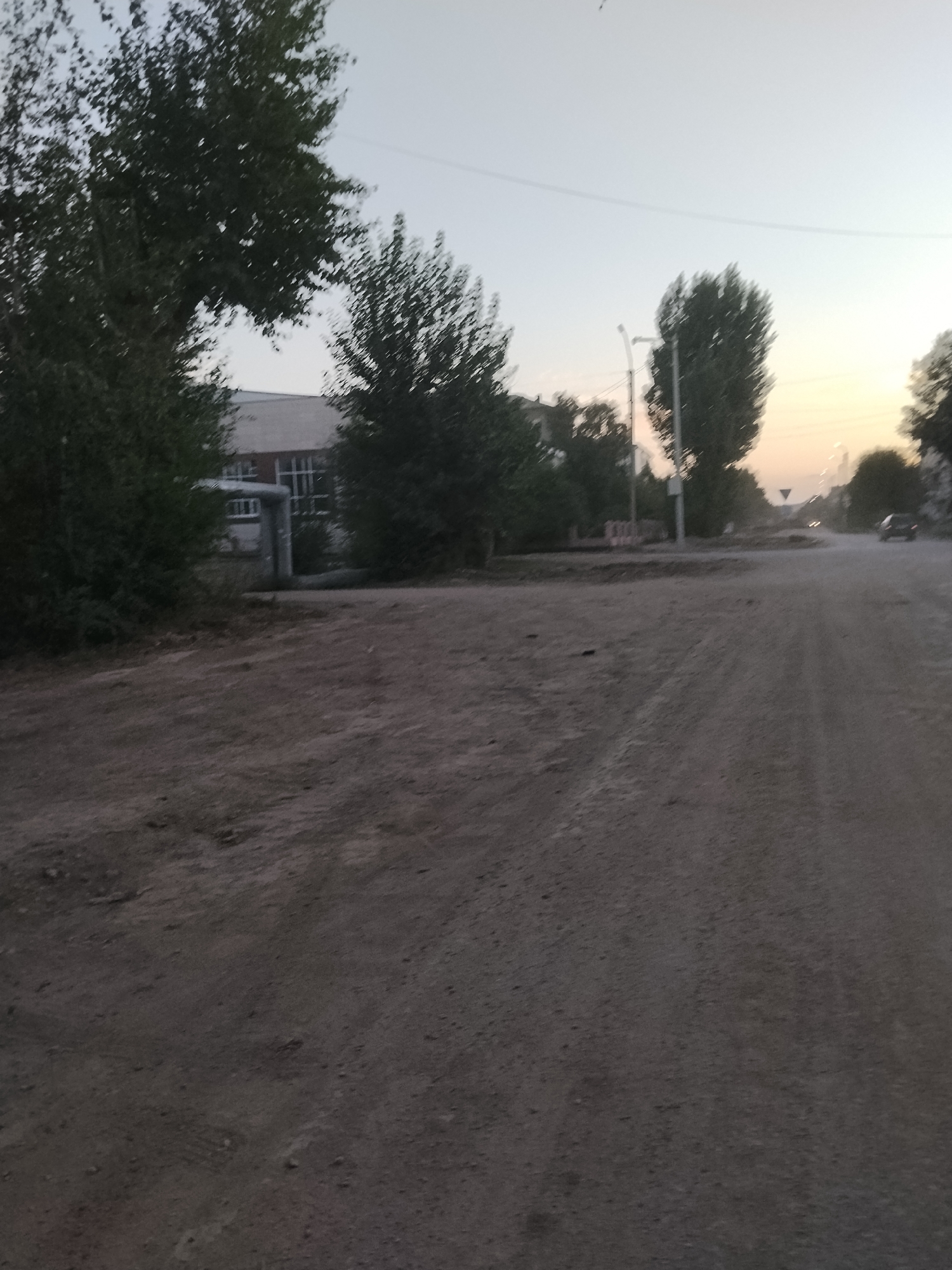 The image depicts a dusty, unpaved road in Kokshetau, Kazakhstan, under a hazy sky. On the left, a building with a white facade and dark windows is partially obscured by dense foliage. Adjacent to the building, a large, grey industrial pipe runs horizontally. Several trees with dark green leaves are visible throughout the scene, some framing the building and others lining the road. A tall, slender lamppost stands near the building. The road itself is made of dirt and gravel, with visible tire tracks. In the distance, the road continues, and a black car is driving away from the viewer. The overall atmosphere is one of a quiet, perhaps rural or industrial, area on a clear but dusty day.
