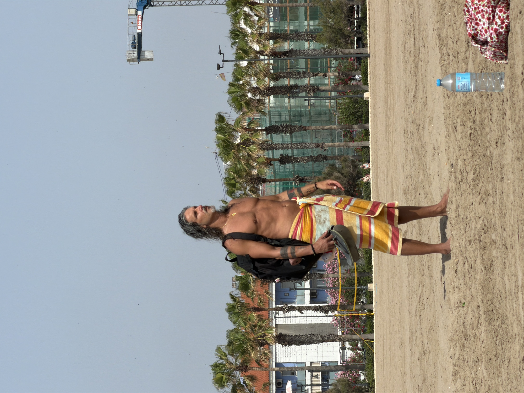 A man with long, dark hair and a beard stands on a sandy beach, facing right. He is wearing a colorful striped sarong and a black backpack slung over his left shoulder. In his right hand, he holds a pair of dark sandals. He has tattoos on his arms and a cross necklace. Behind him, palm trees line the shore, with buildings and a construction crane visible in the distance. A yellow volleyball net stands in the middle ground. A plastic bottle of water and a patterned cloth lie on the sand to the right of the man. The sky is clear and pale blue.
