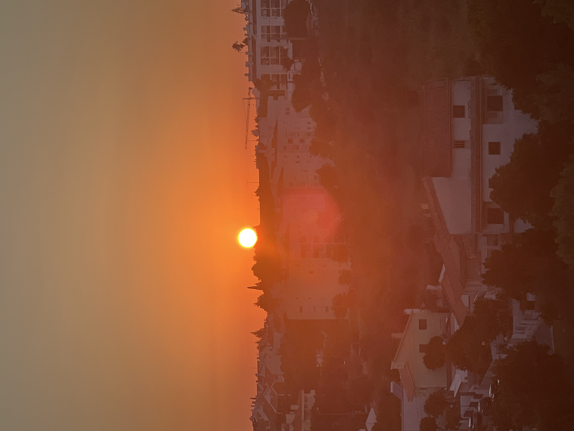 This is a photograph of a sunset over the city of Cascais, Portugal. The sun is a bright, orange orb low in the sky, casting a warm glow over the scene. In the foreground, there are several residential buildings with red-tiled roofs and white facades. Trees and vegetation are interspersed throughout the residential areas. In the mid-ground and background, more buildings, including apartment complexes and some under construction with a crane, are visible. The sky transitions from a bright orange near the horizon to a softer, lighter orange and then to a pale yellow-orange at the top of the frame. The overall impression is one of a peaceful evening in a coastal European city.